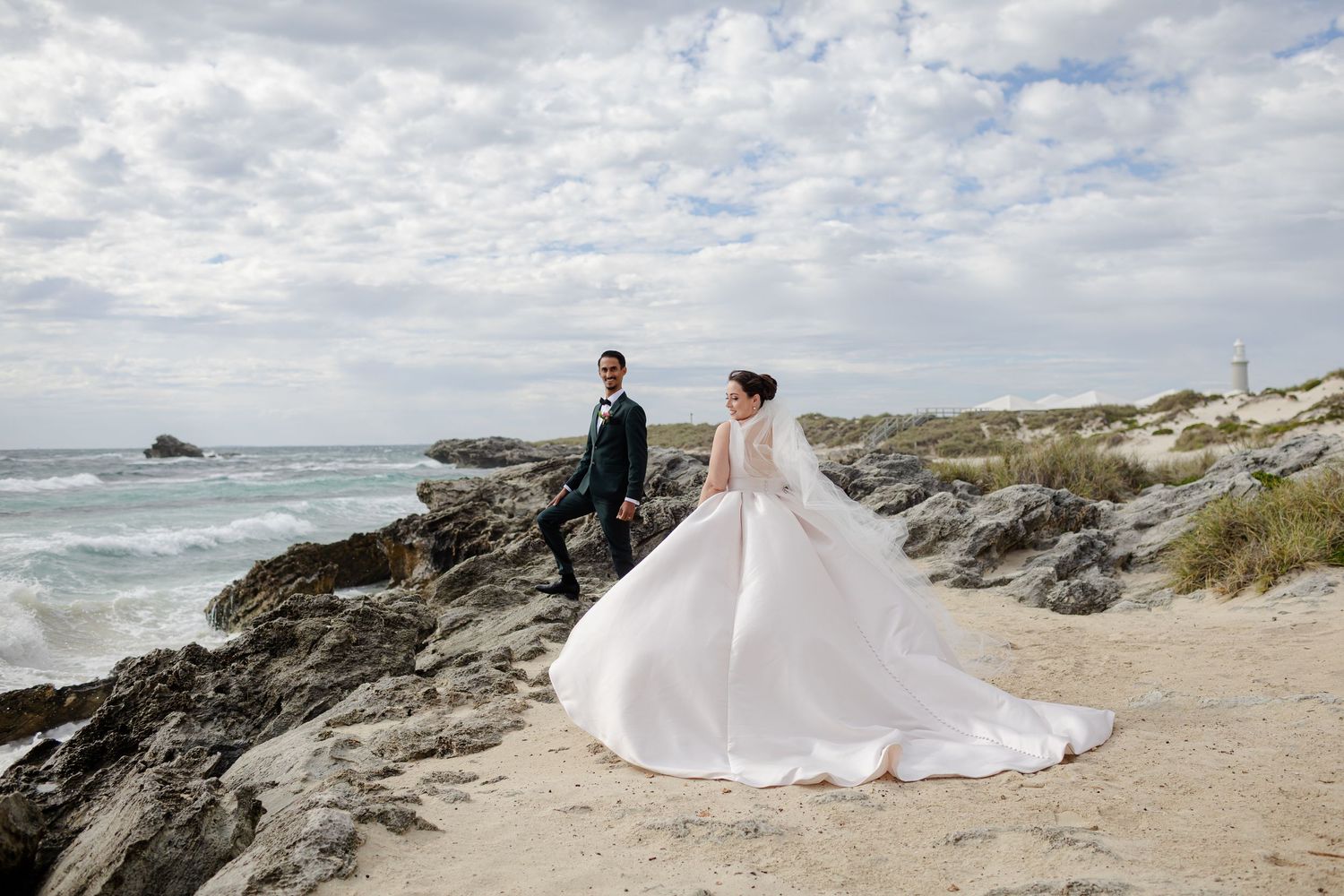 A couple in wedding attire stands on a rocky beach coastline under cloudy skies in Cornwall.