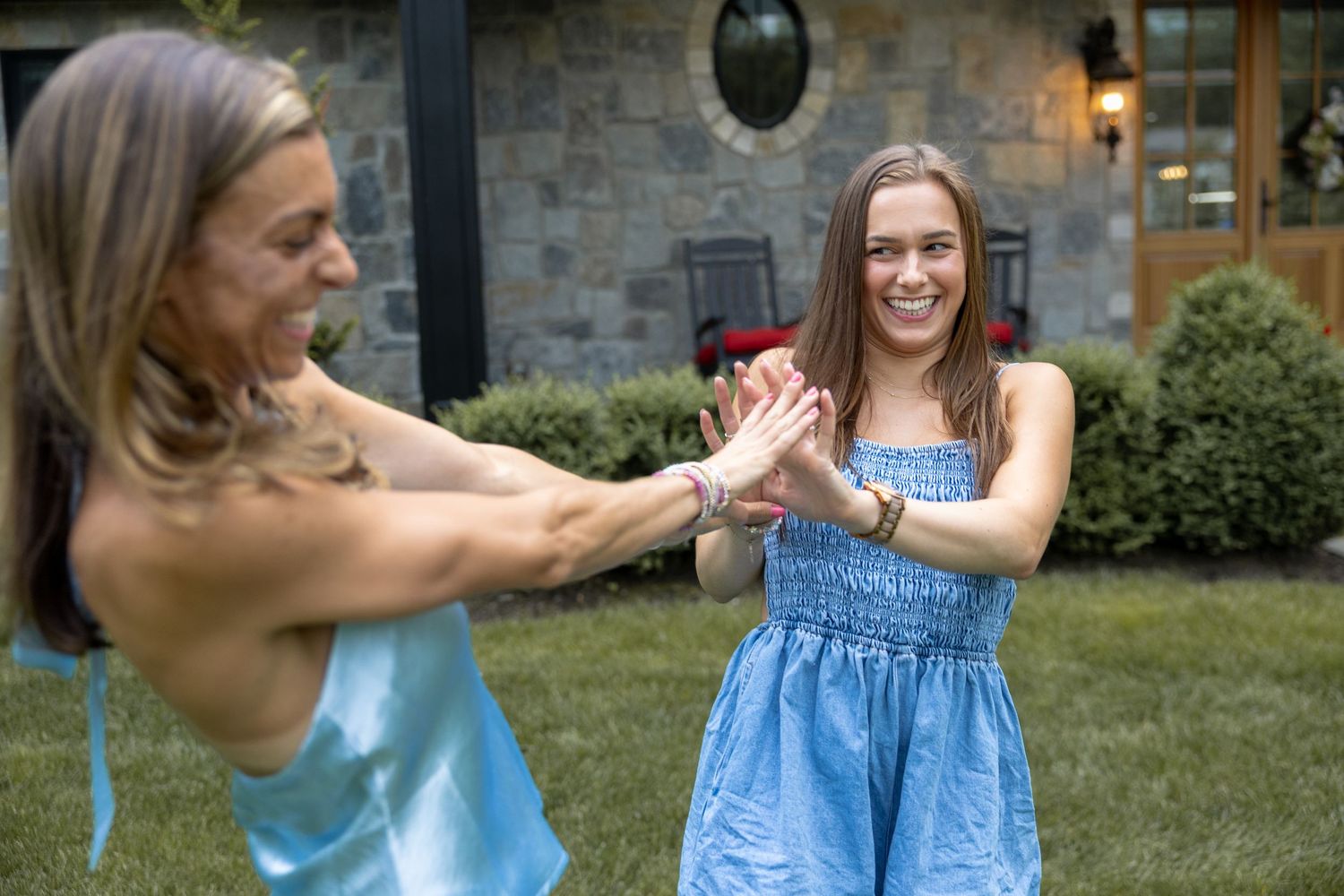 Two people in blue dresses play a summertime game outside a stone house with garden shrubs.