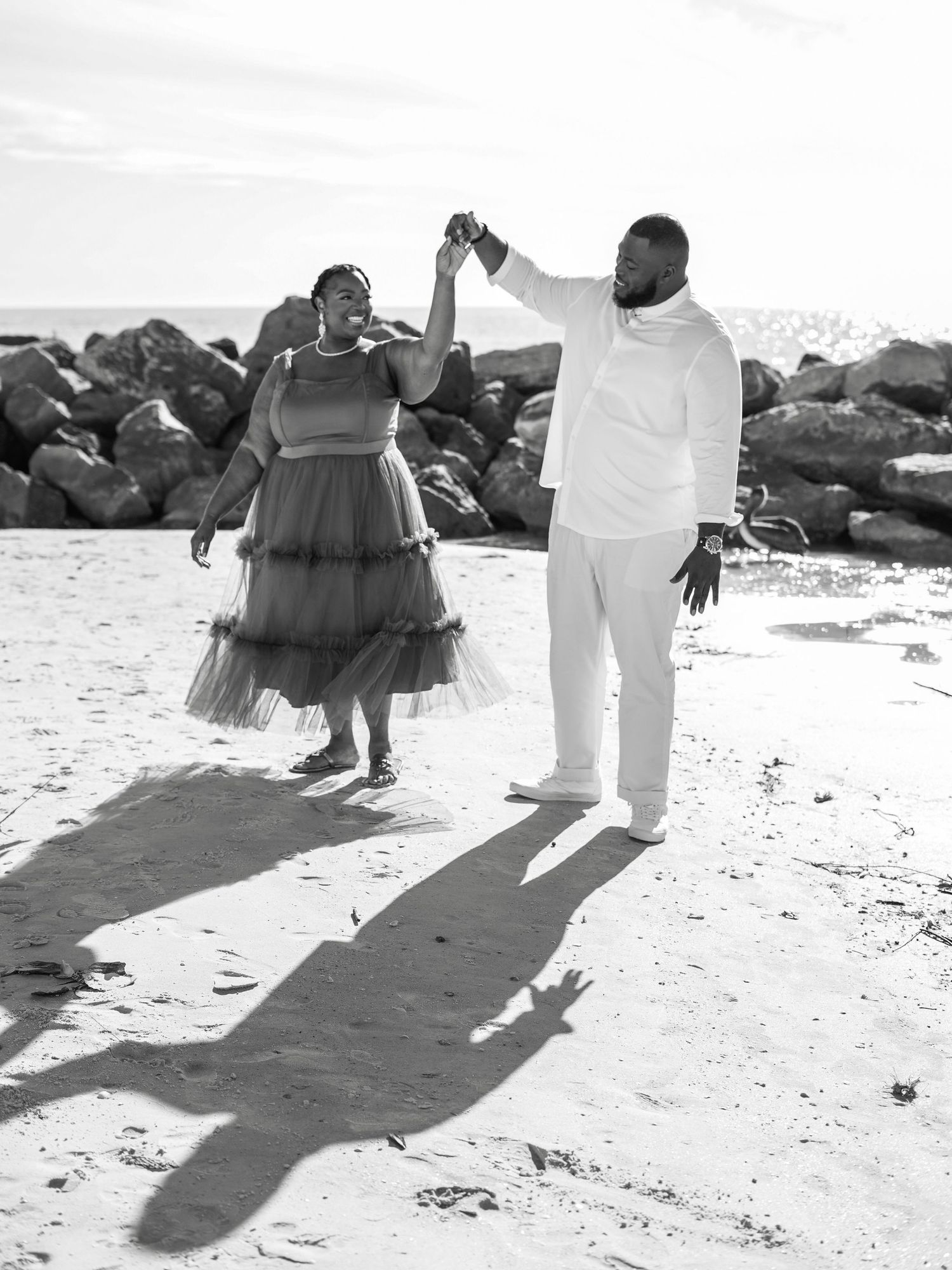 A romantic black and white photo of a couple dancing together on a beach by some rocks, casting long shadows at sunset.