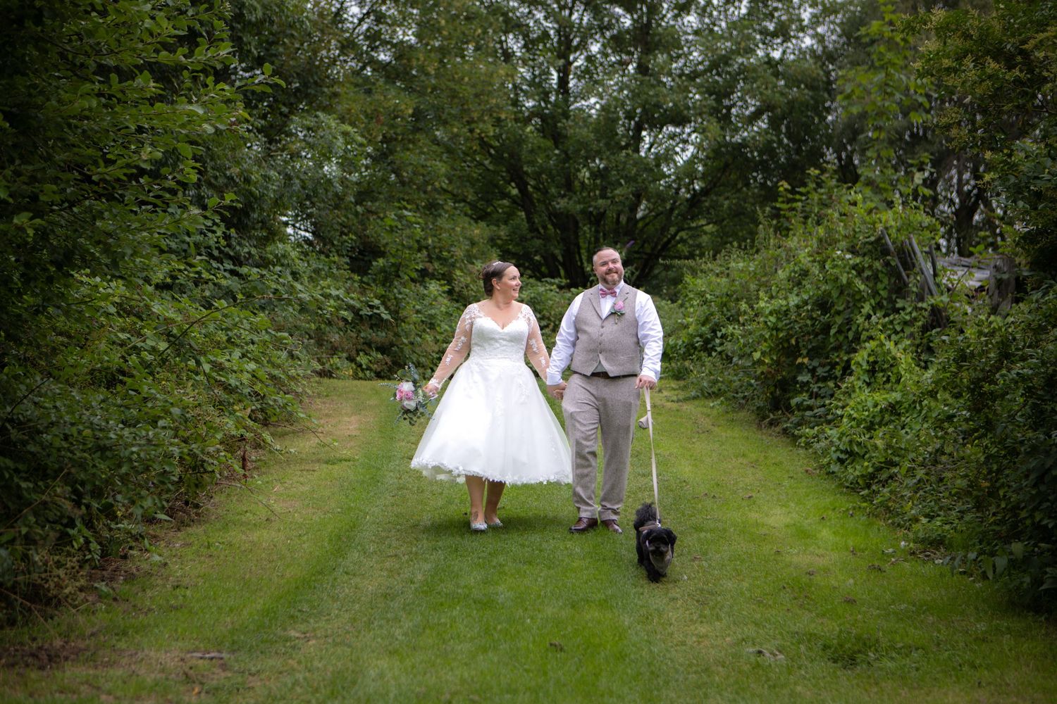 A couple walks together with their black dog down a green path surrounded by lush summer foliage.