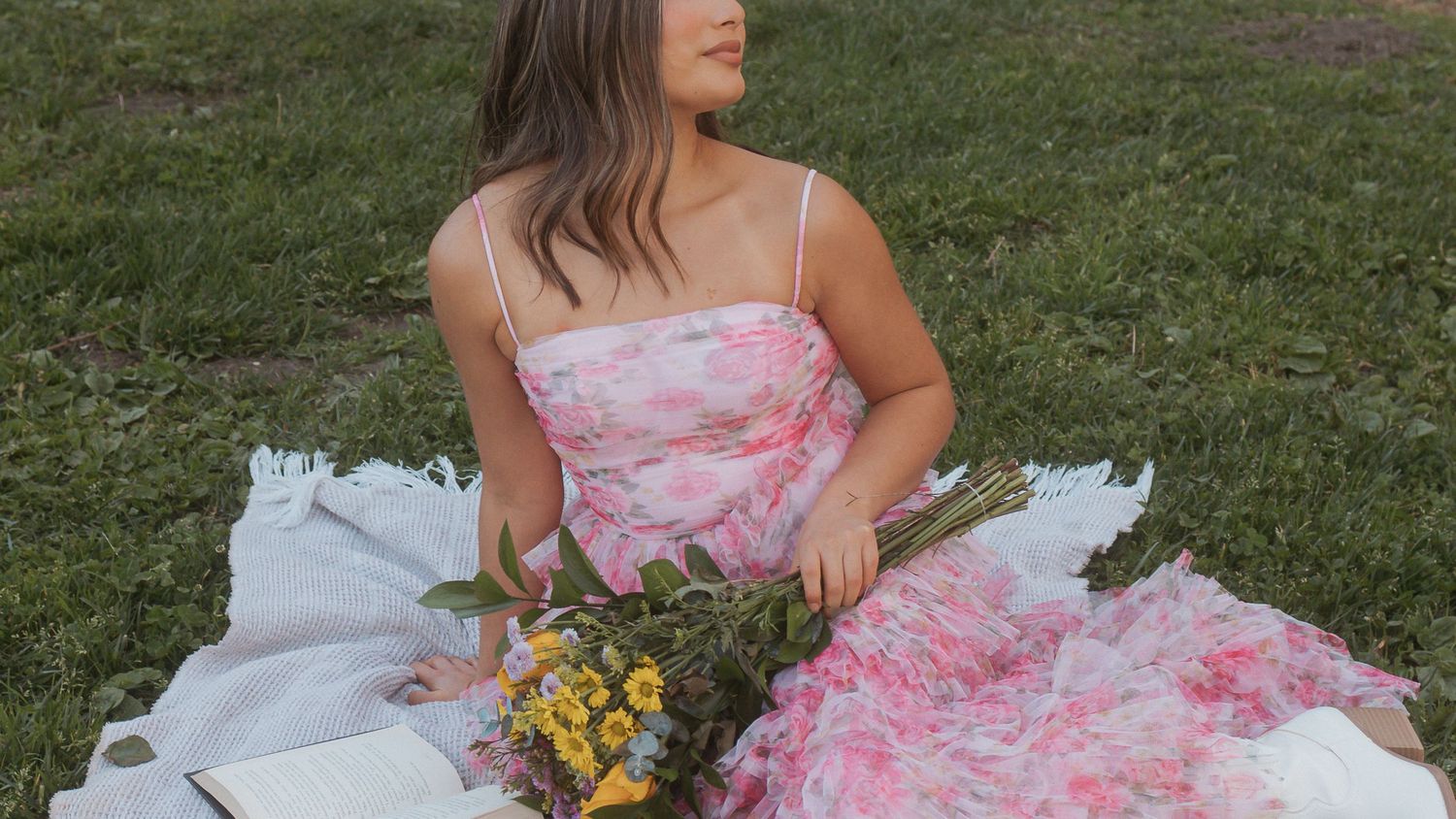 A person in a pink dress sits on white fabric in a grassy field with scattered yellow flowers.