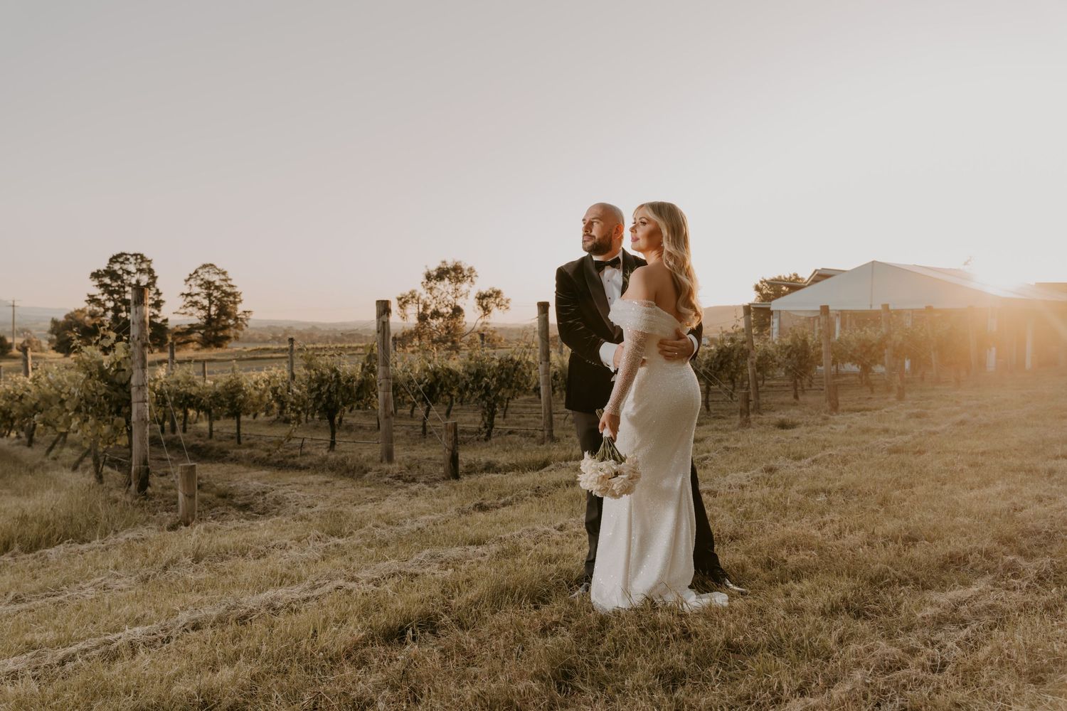 A couple in wedding attire embraces at sunset on a grassy hill overlooking the countryside.