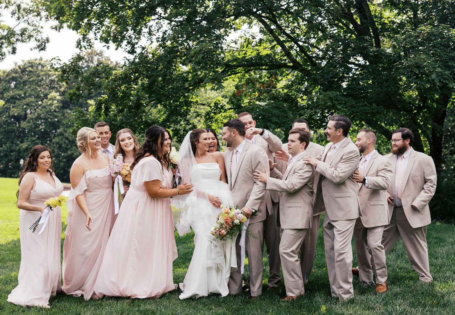 Wedding party in blush pink dresses and light beige suits pose together under green trees on sunny afternoon.