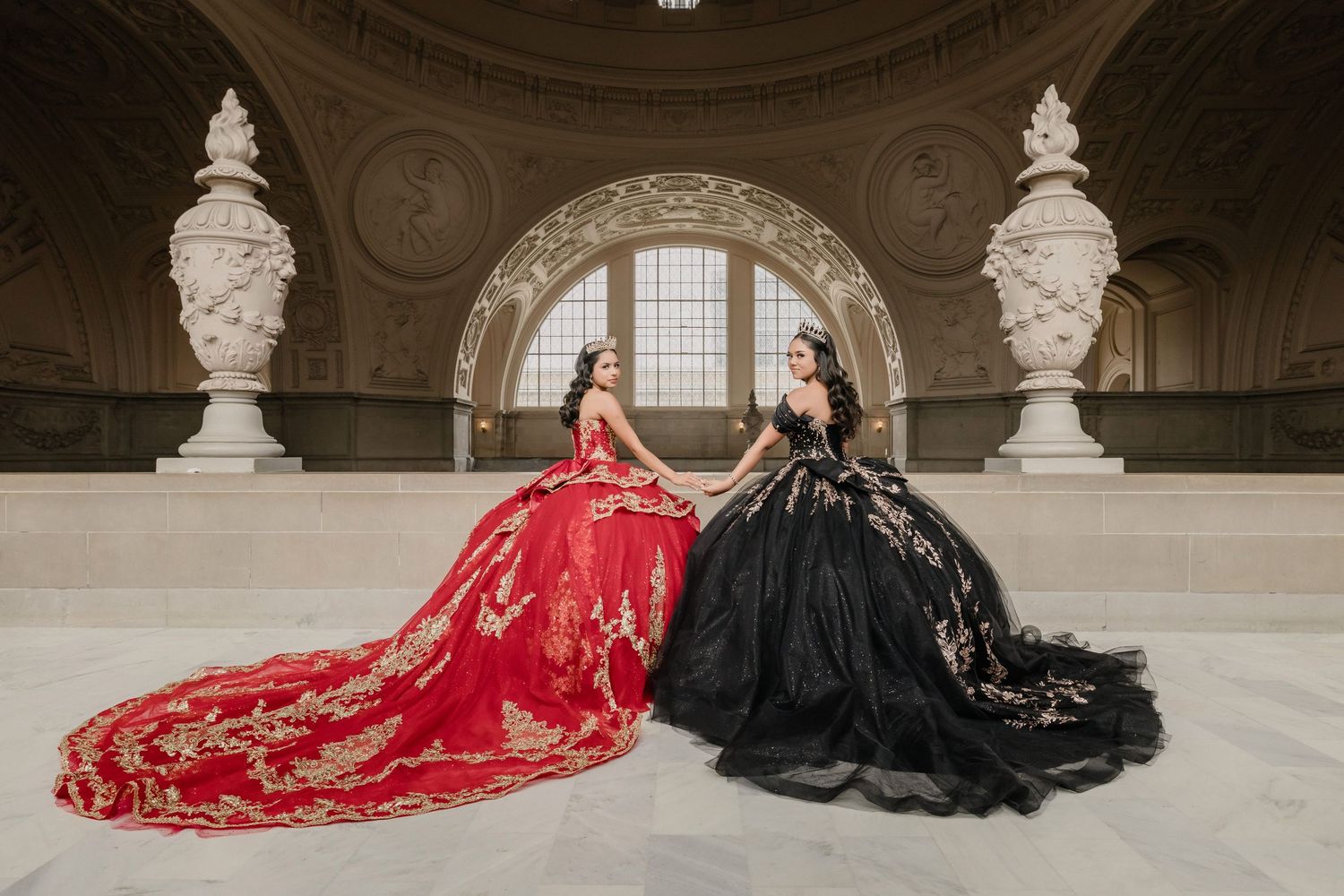 Two quinceañeras in dramatic red and black gowns pose elegantly in a grand historic ballroom with ornate architecture.