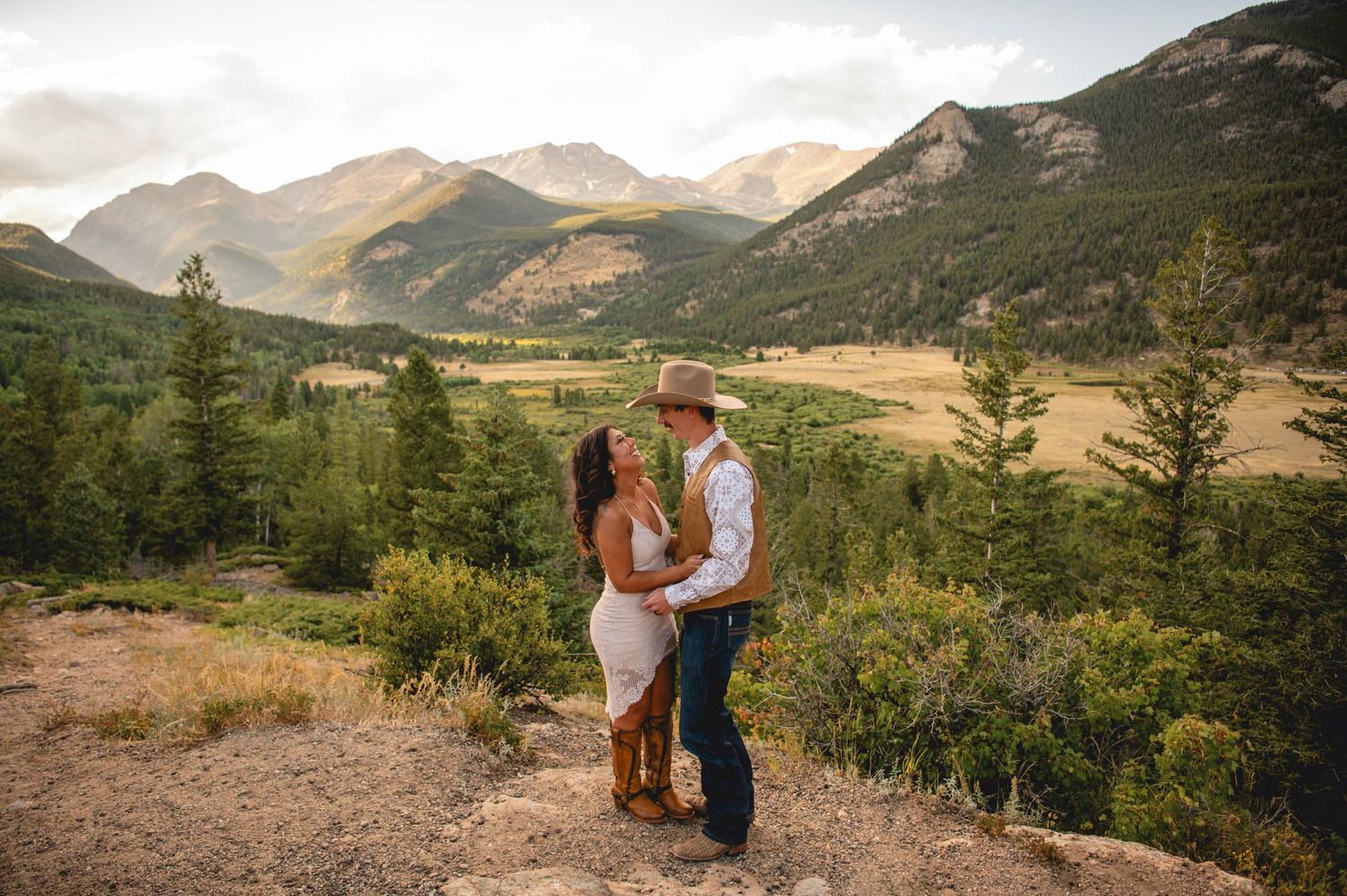 Two people embrace in a romantic pose against a backdrop of majestic mountains and scenic wilderness landscapes.