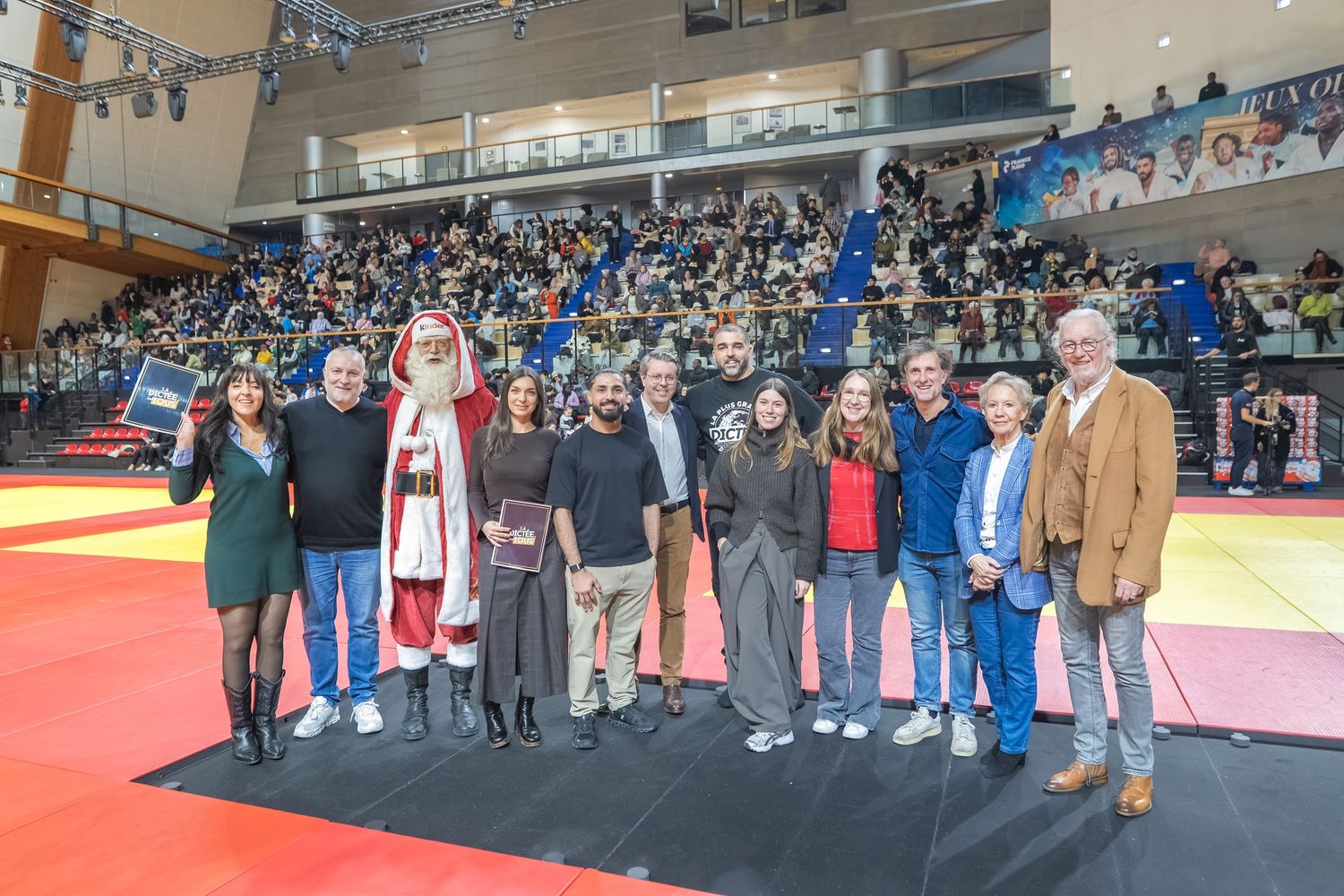 Group of people standing together at a martial arts or judo competition venue with red and yellow mats visible.