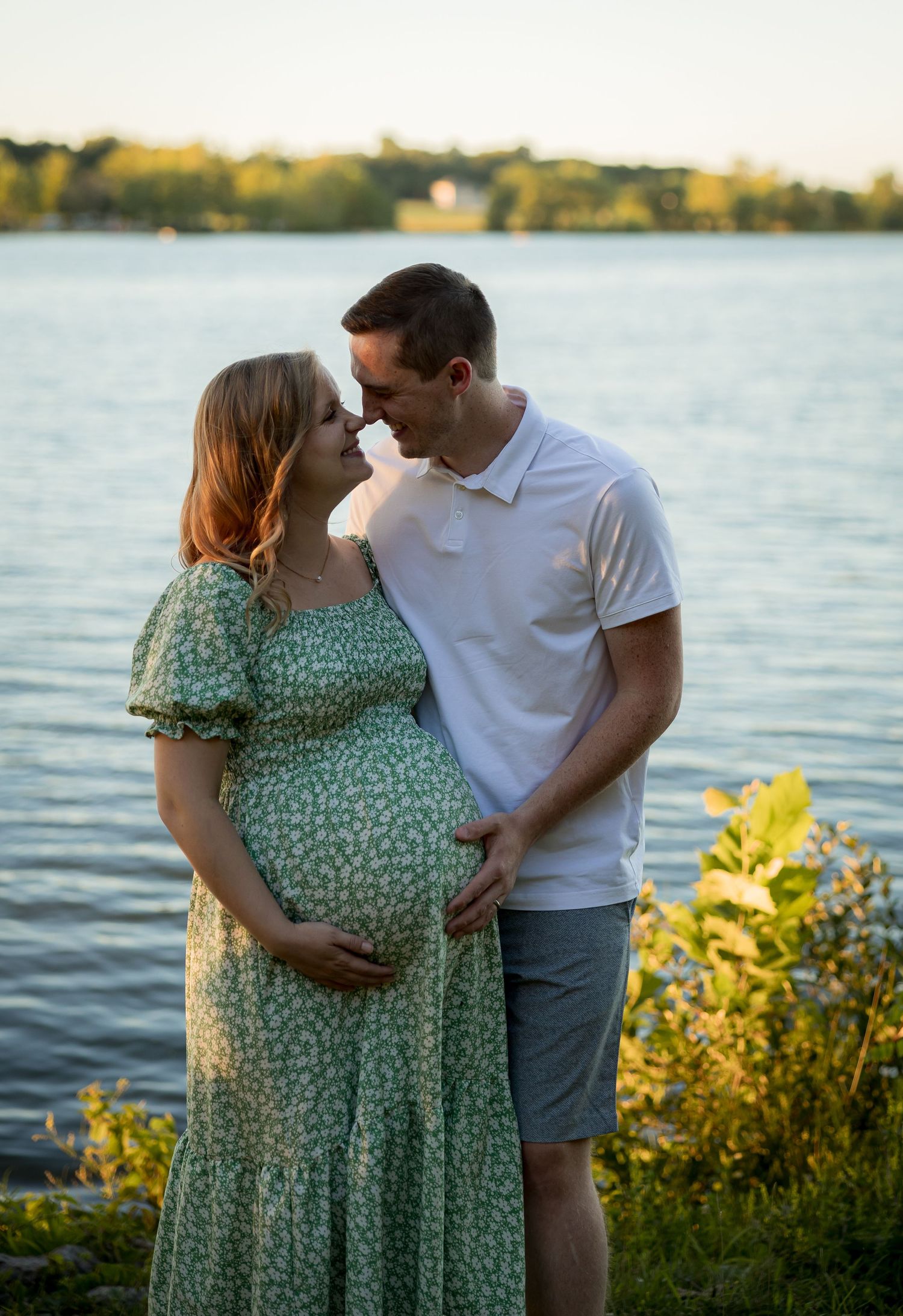 Waterfront maternity session with couple in casual attire against blue water and evening sky.