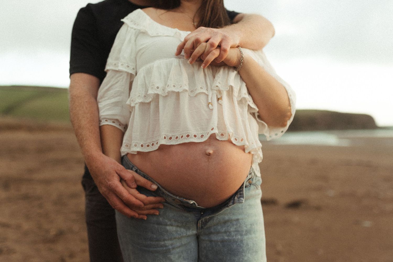 A pregnant woman in a white lace top poses at a sandy beach during sunset.