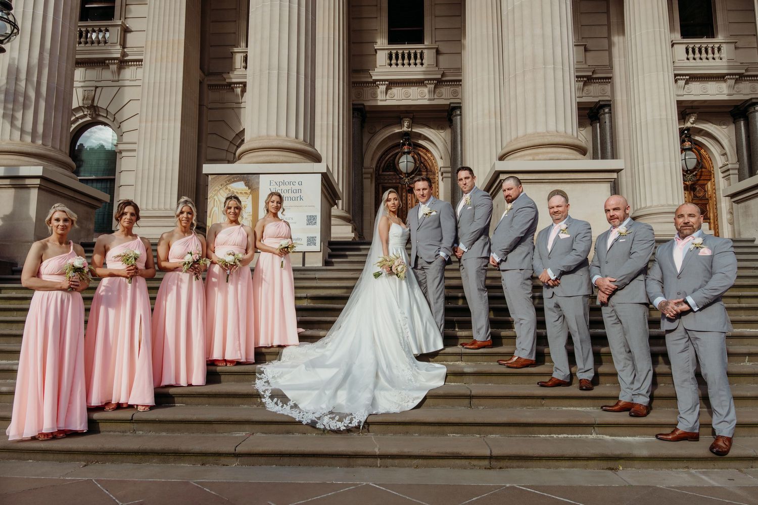Wedding party in pink bridesmaid dresses and gray suits poses on grand stone steps of historic building with columns.