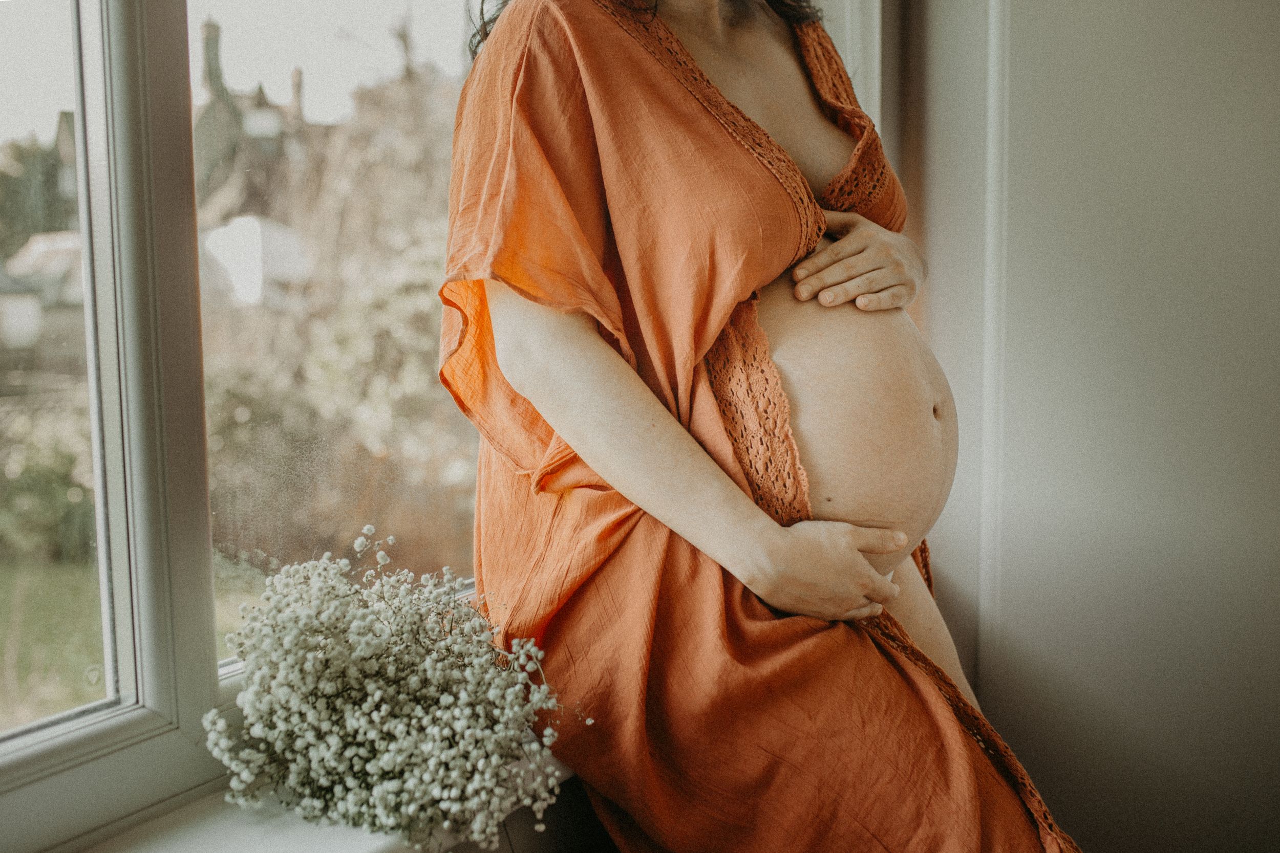 Maternity photo series in warm orange tones showing a pregnant woman's belly and baby's breath flowers by a window.
