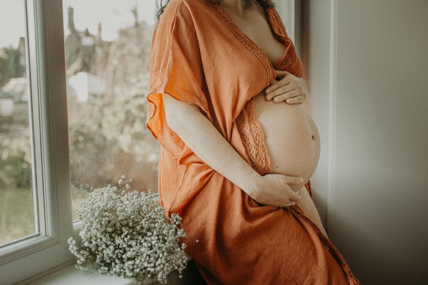 Maternity photo series in warm orange tones showing a pregnant woman's belly and baby's breath flowers by a window.