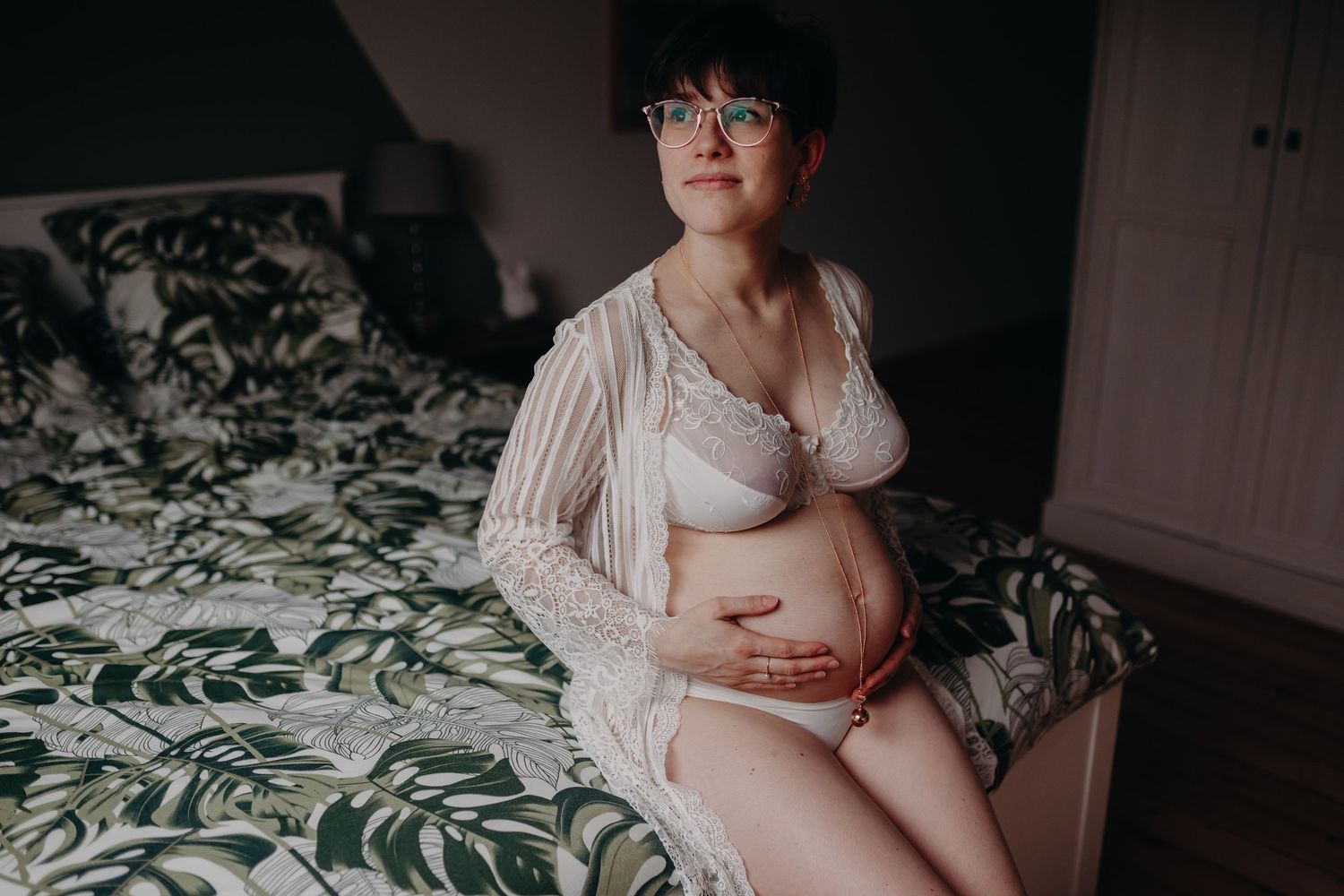 Pregnant woman with short hair and glasses sitting on bed in white lace lingerie.
