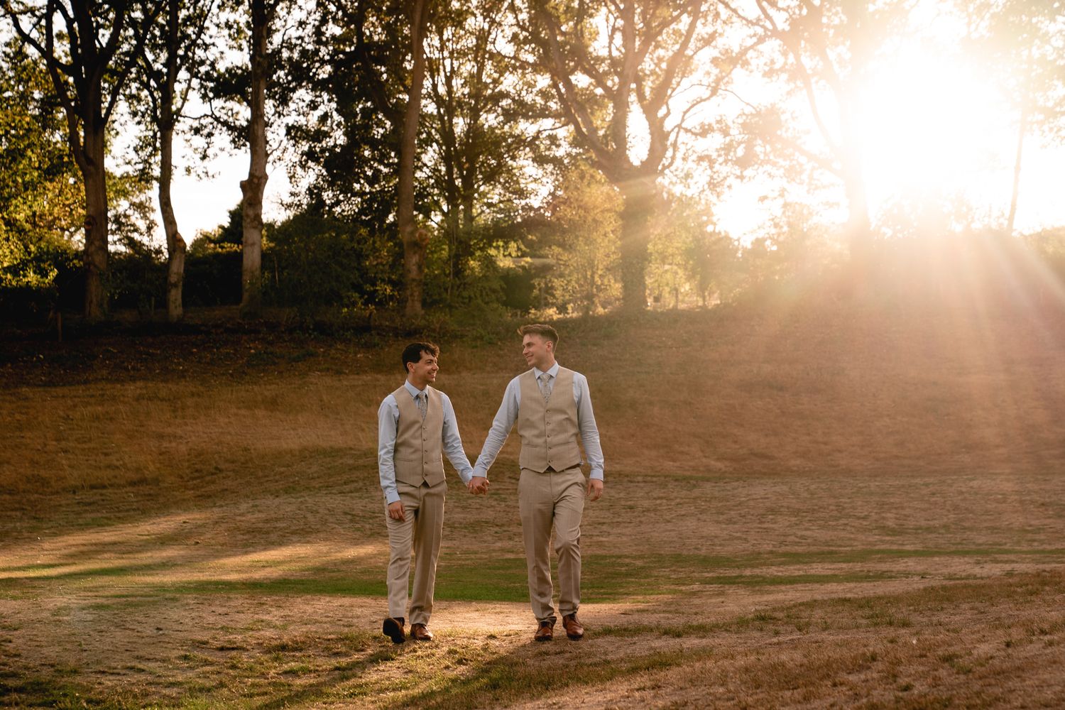 Two grooms in matching tan suits hold hands walking at sunset in a golden outdoor wedding portrait.