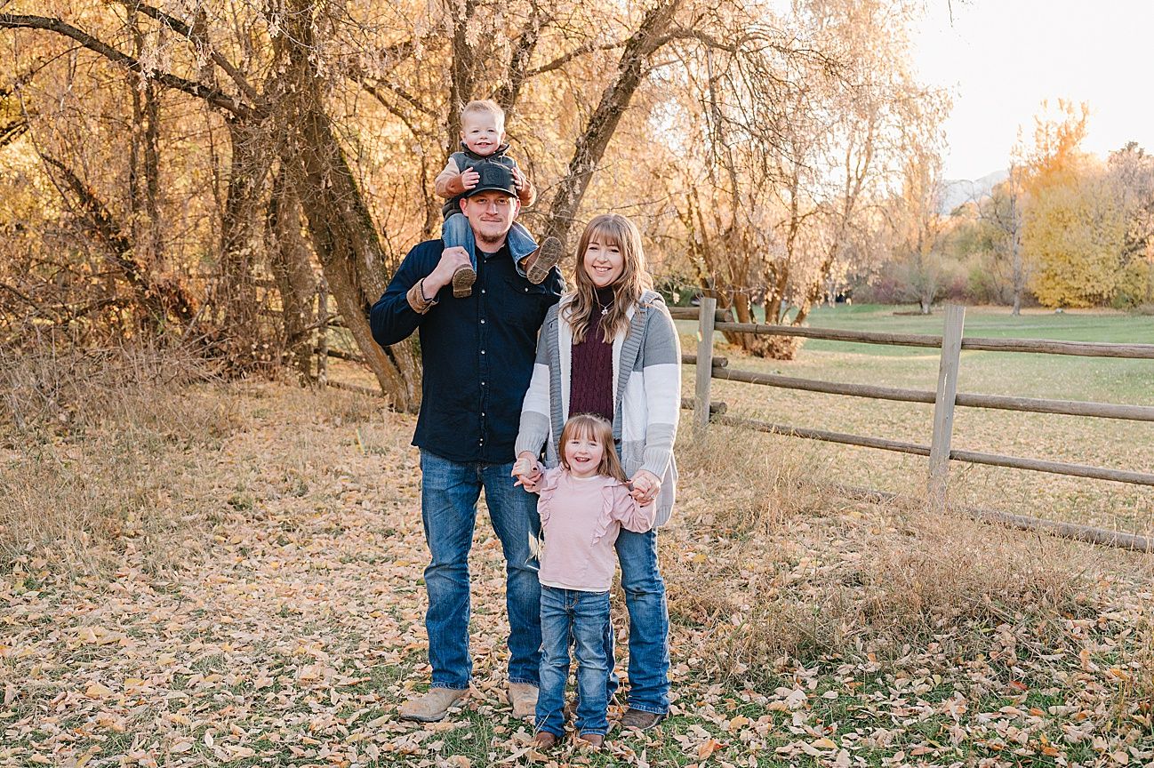 A family stands together on an autumn day with fallen leaves and a wooden fence in the background.