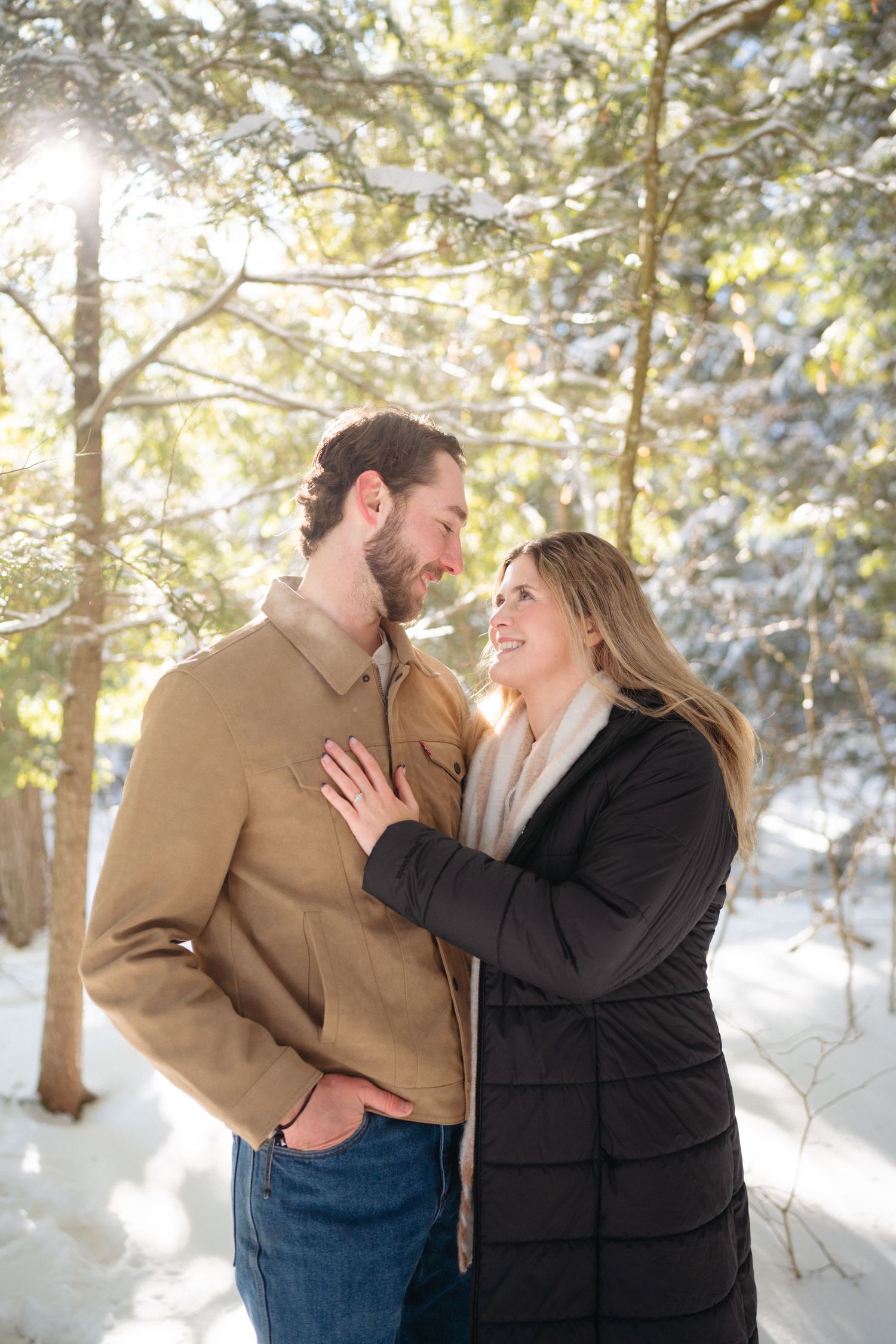 A couple shares a joyful moment together in a snowy winter forest with sunlight streaming through the trees.