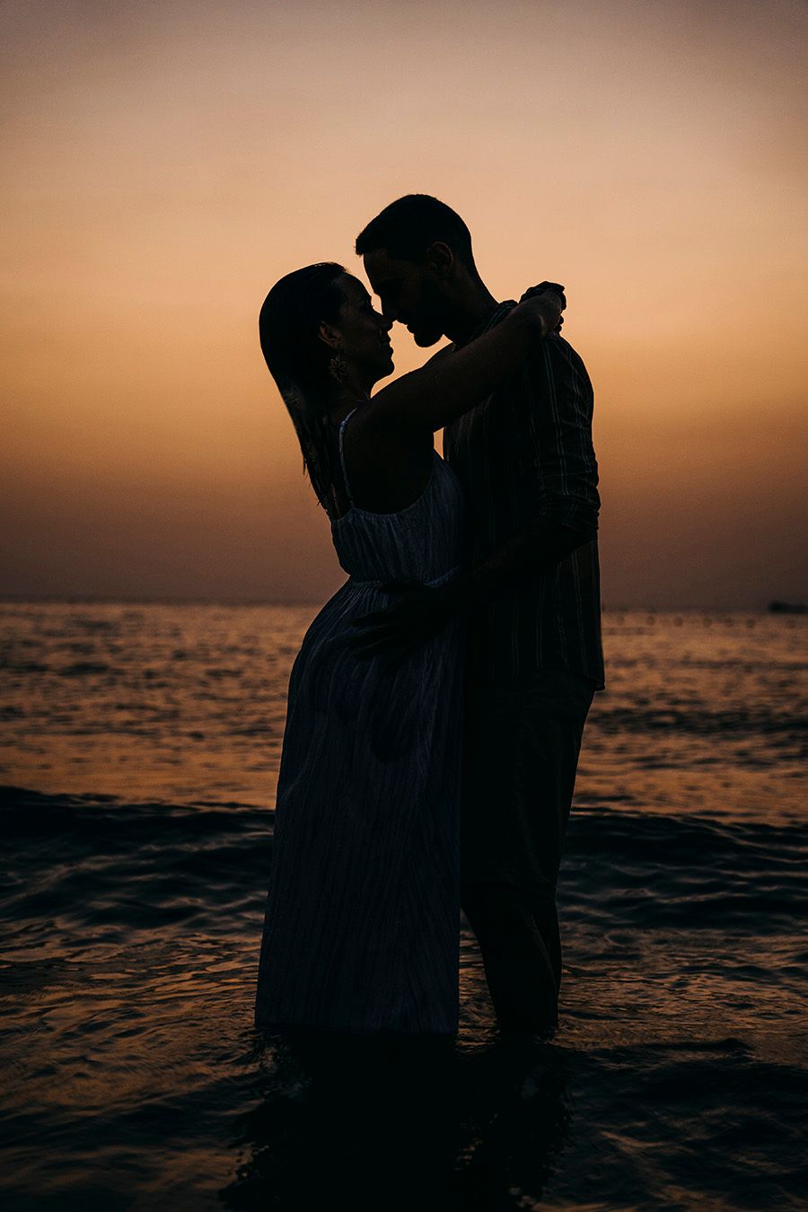 Romantic silhouette of a couple embracing in ocean water at sunset with golden orange sky backdrop.