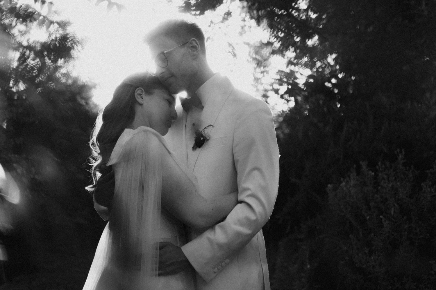 Black and white wedding photo of bride and groom embracing outdoors, backlit by sunlight with lush greenery behind them.
