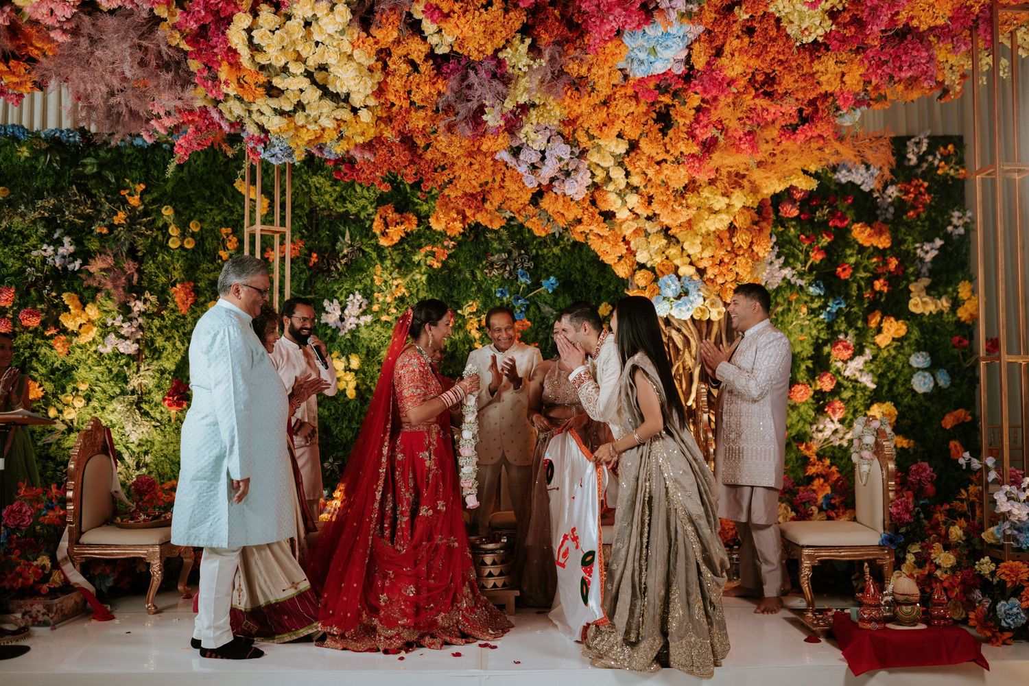 Colorful Indian wedding ceremony under vibrant floral canopy with bride in red lehenga and guests in traditional attire.