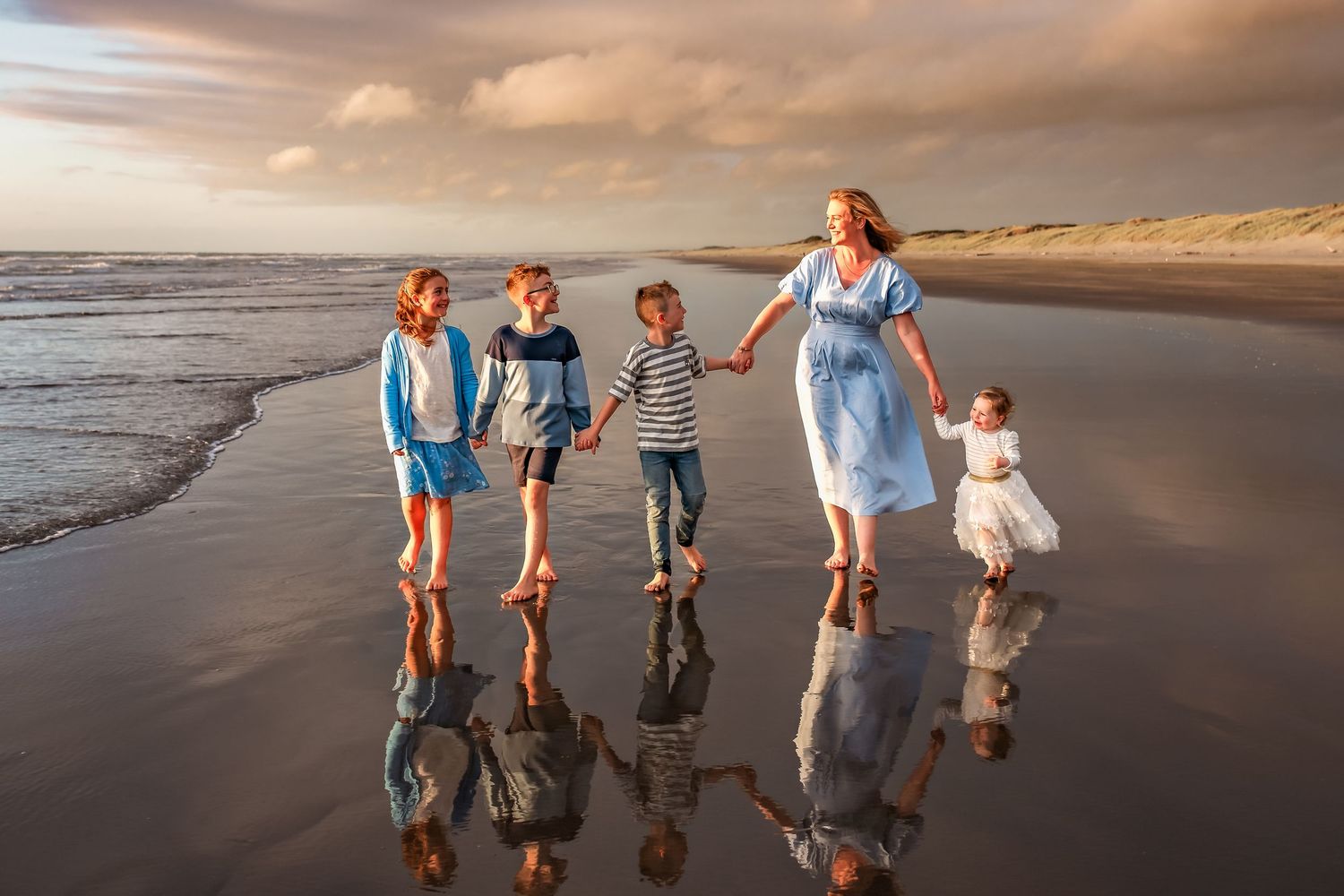 A group walks together on a reflective beach at sunset, their silhouettes mirrored in the wet sand along the shoreline.