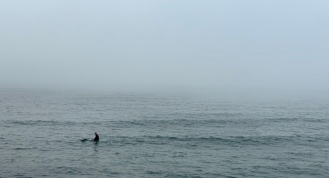 A surfer rides a wave in foggy conditions on a misty gray ocean morning.