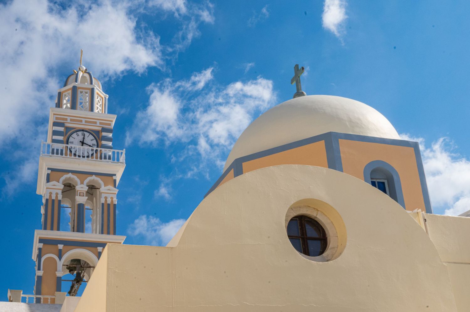 A white Mediterranean church with a bell tower and dome against a bright blue sky with puffy clouds in Santorini, Greece.