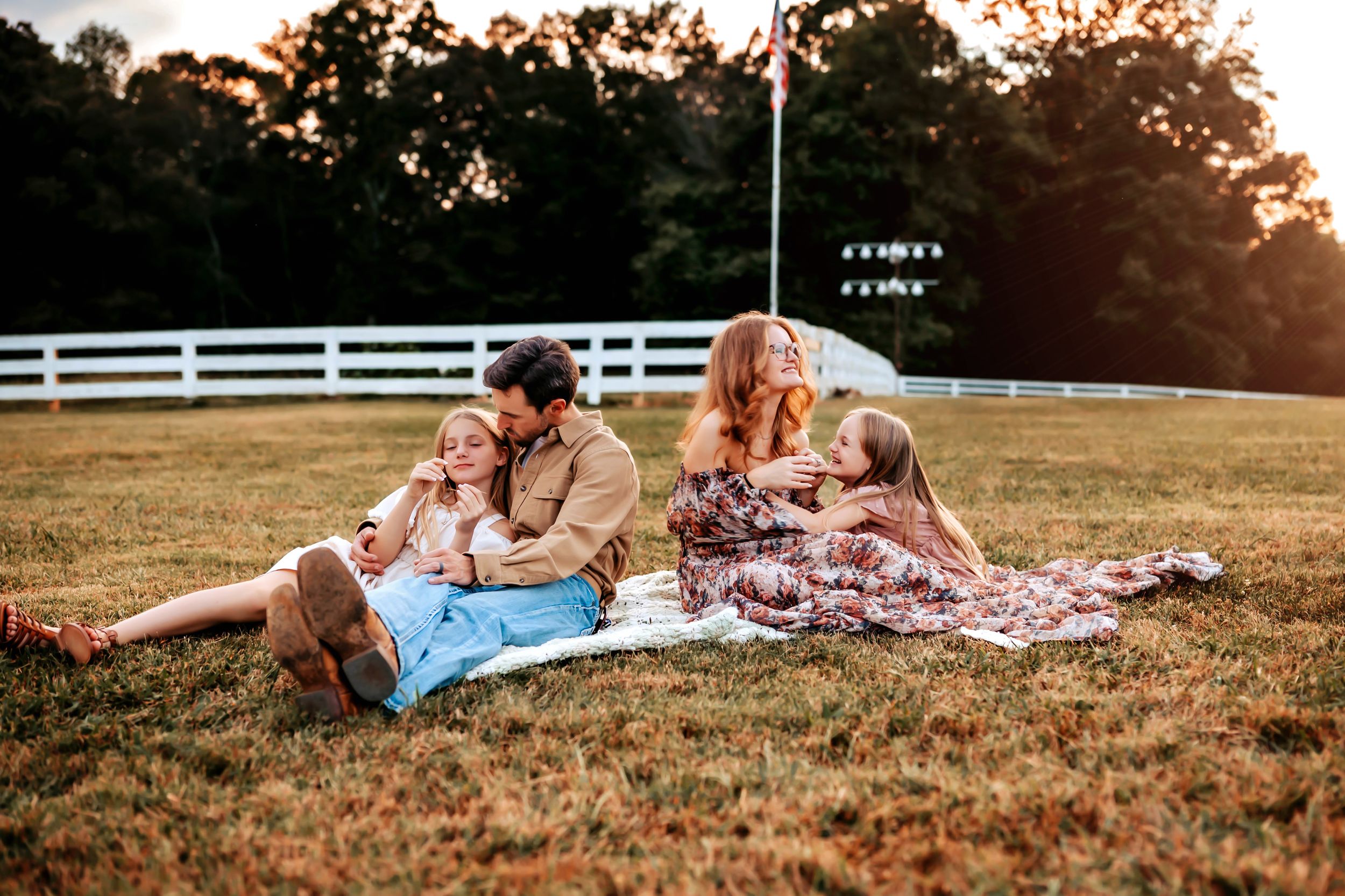 A romantic couple embraces at sunset near a white fence on a rustic farm property.