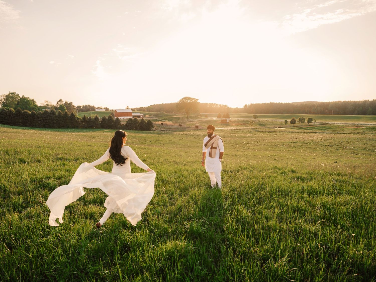 Couple in white attire standing in a sunlit green field at golden hour with rolling hills in the background.