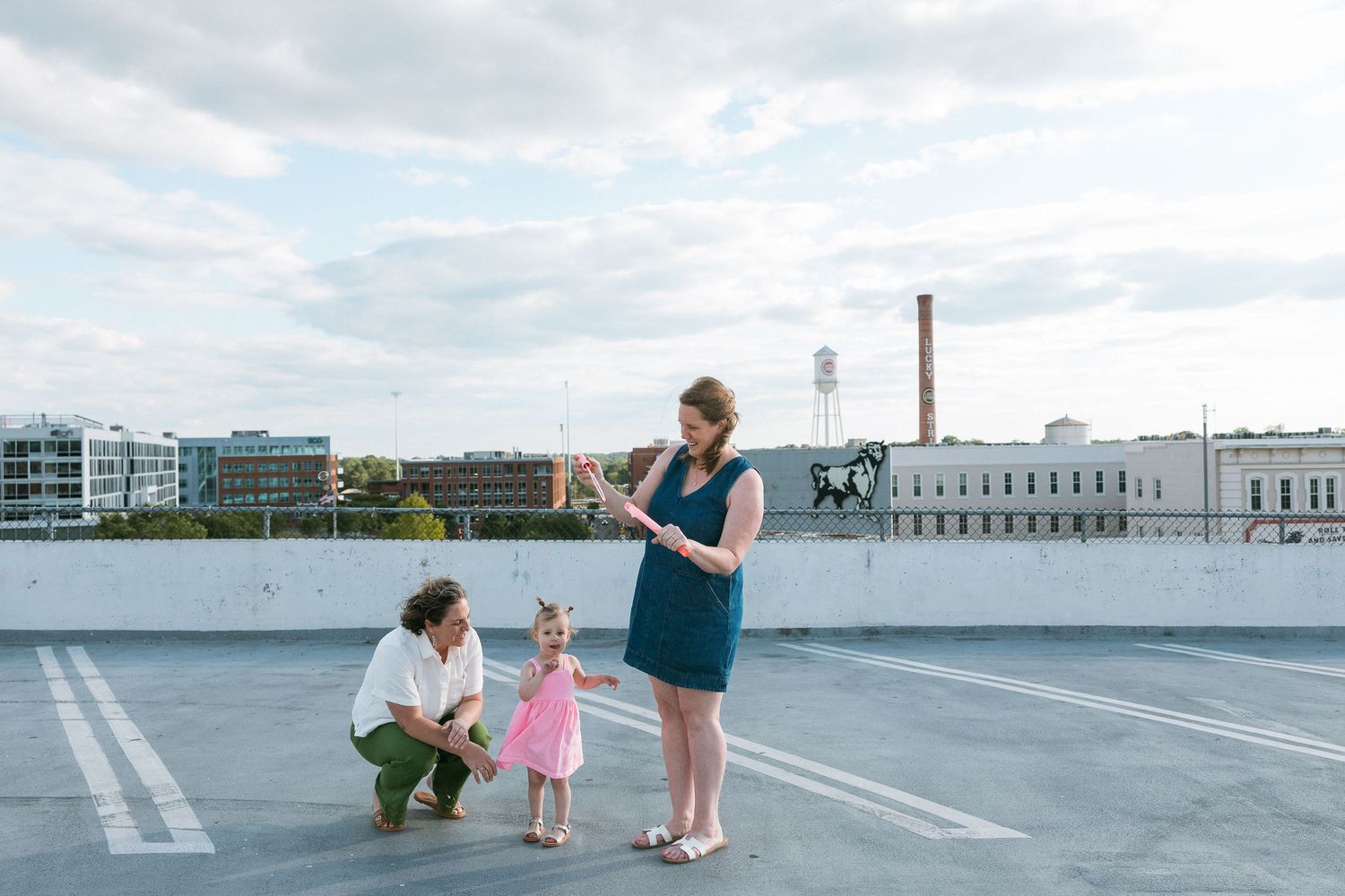 Family of three poses on rooftop parking deck with city skyline, parents with toddler girl in pink dress.