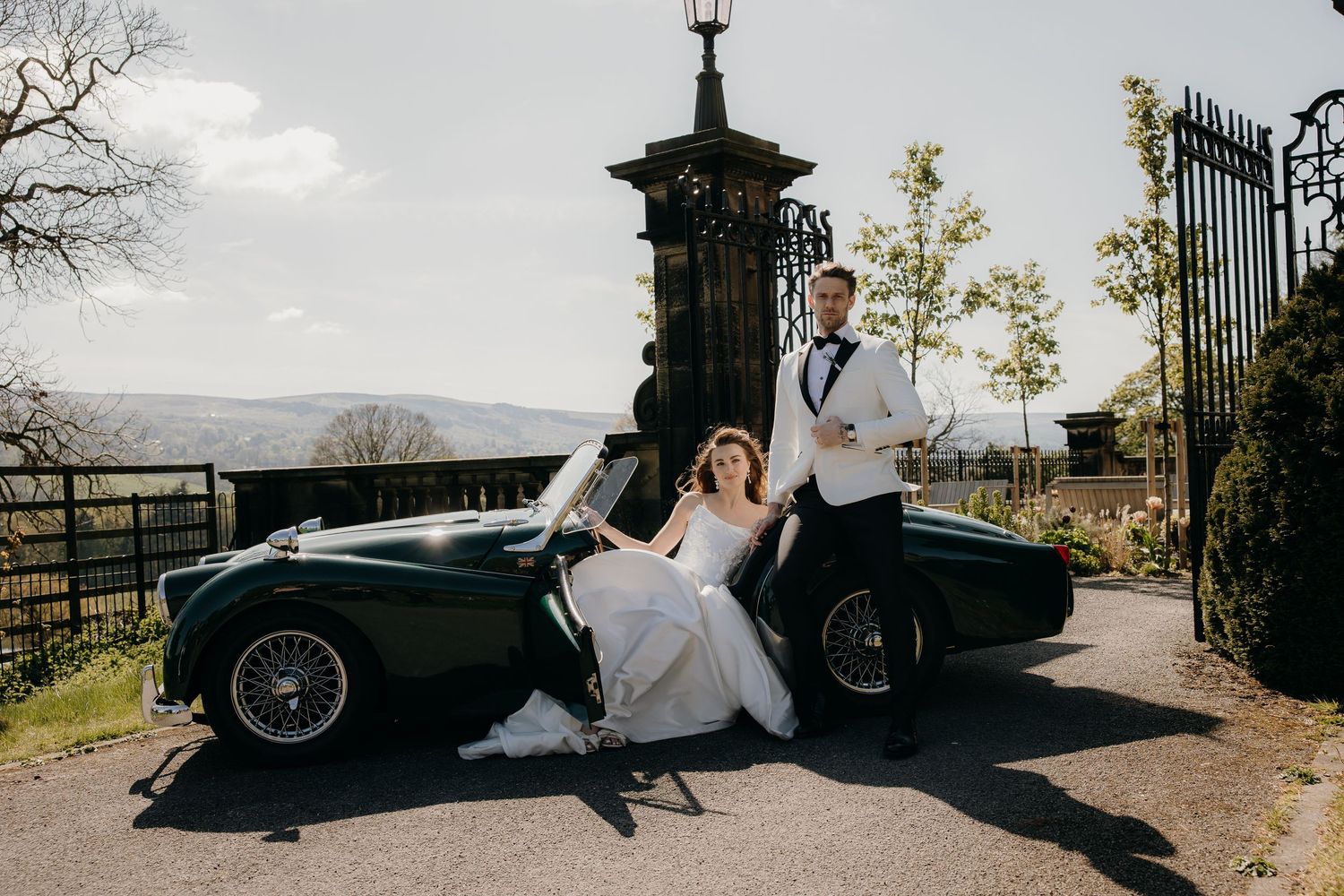 Bride and groom posing with a vintage green sports car at an elegant estate on their wedding day.