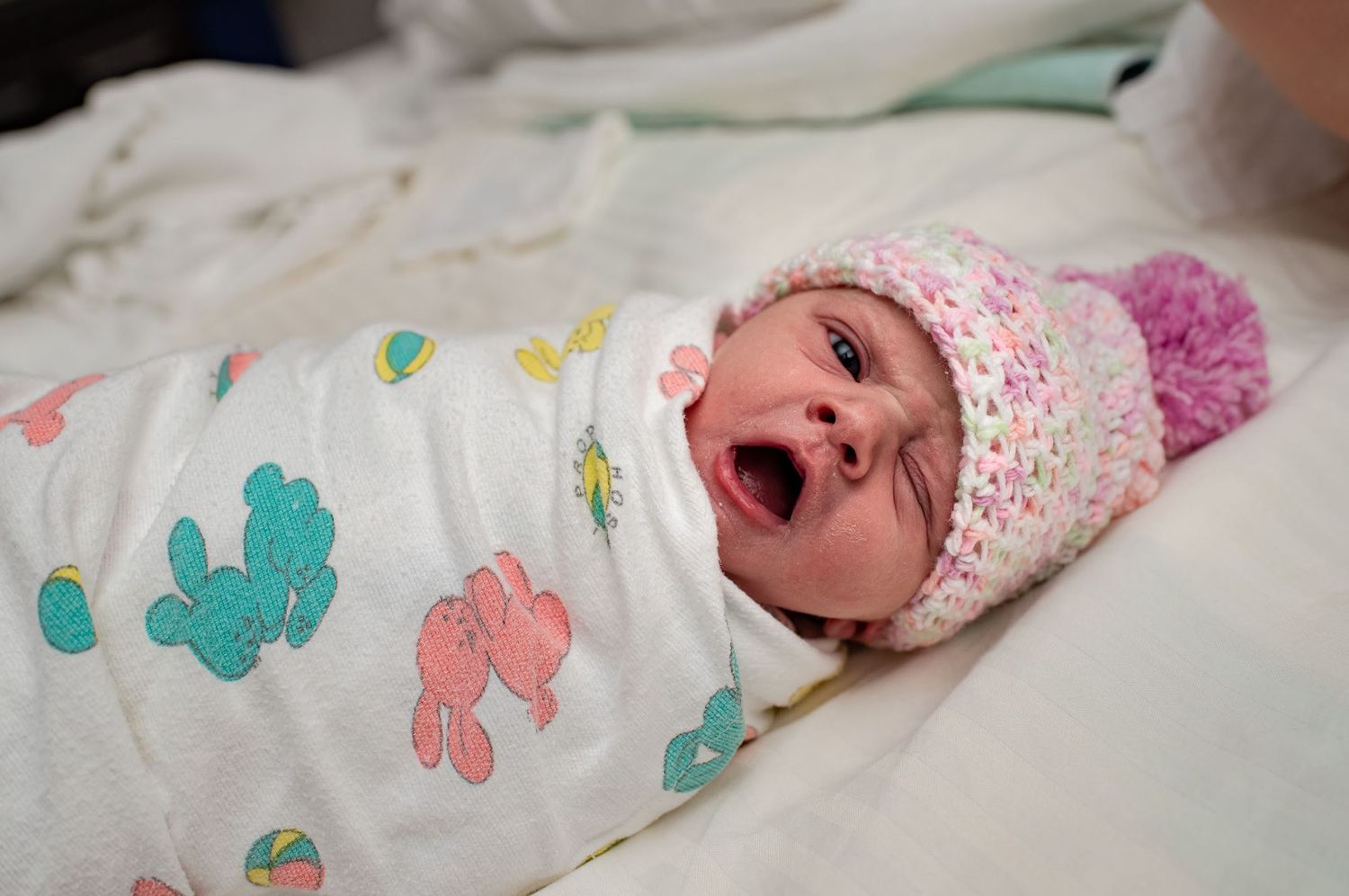 Newborn baby wearing floral swaddle and pink hat yawning.
