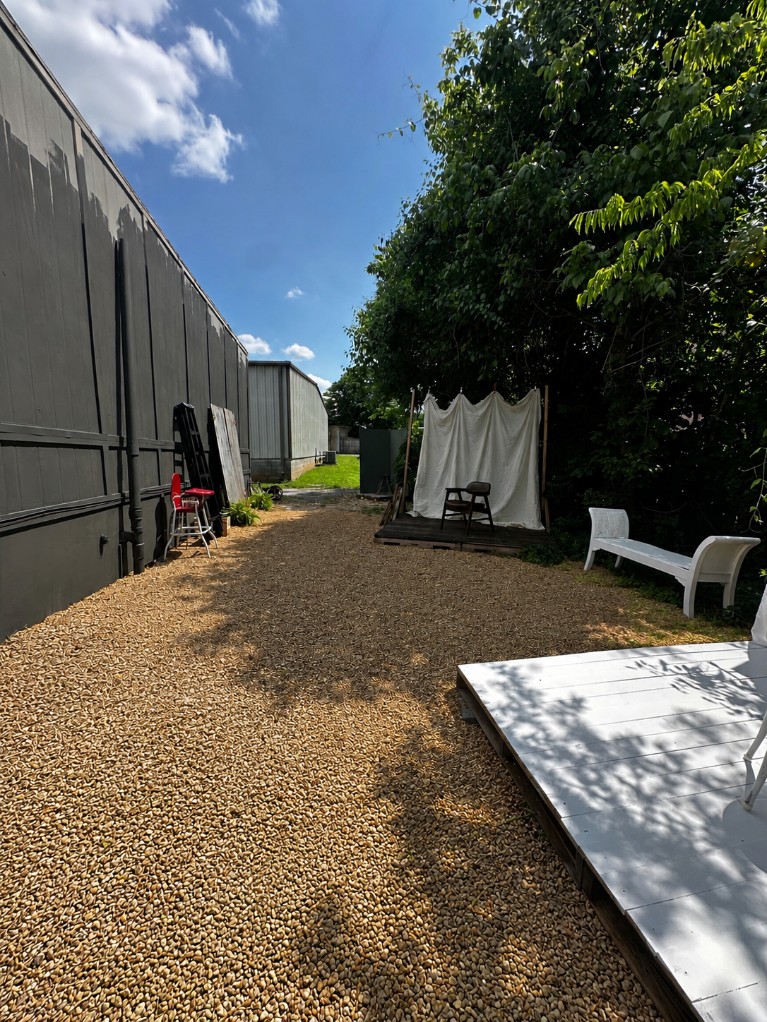 Gravel pathway between white metal buildings and green trees under blue sky with white tent and chairs visible.