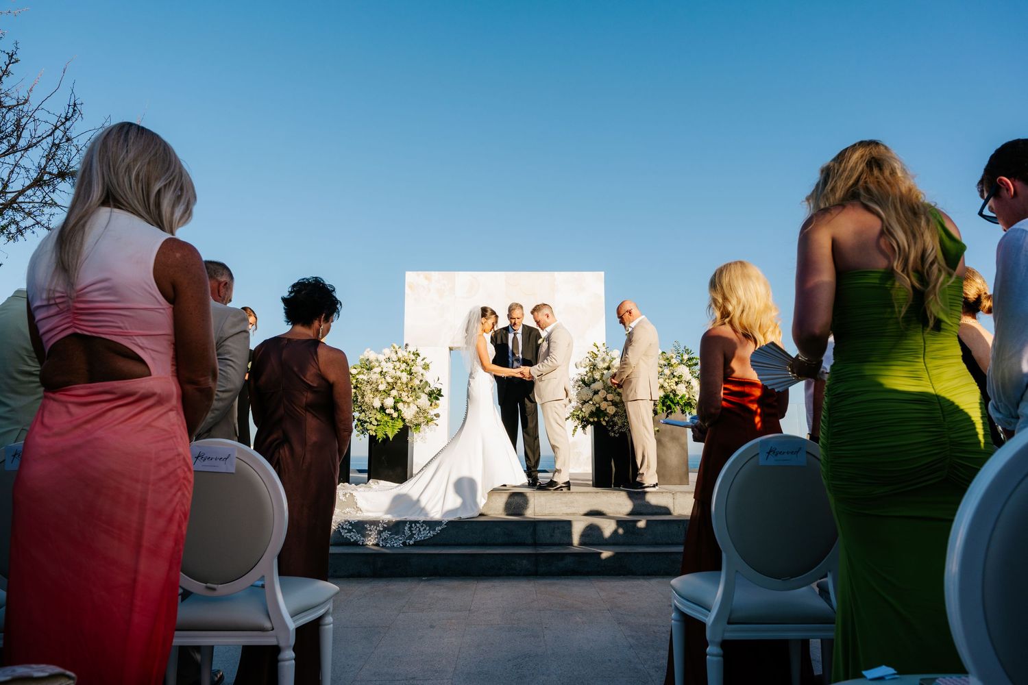 Outdoor wedding ceremony under blue sky with bride and groom at altar, guests in colorful dresses standing in aisle.