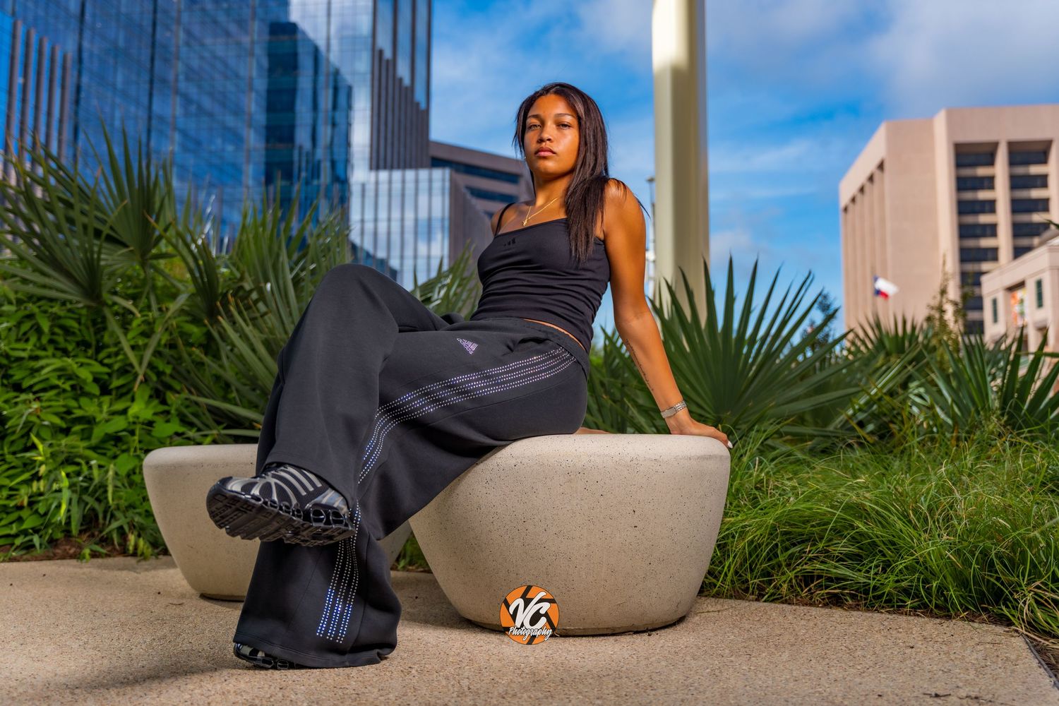 A young Black woman in black Adidas tracksuit sits on a concrete bench surrounded by tropical plants and city buildings.