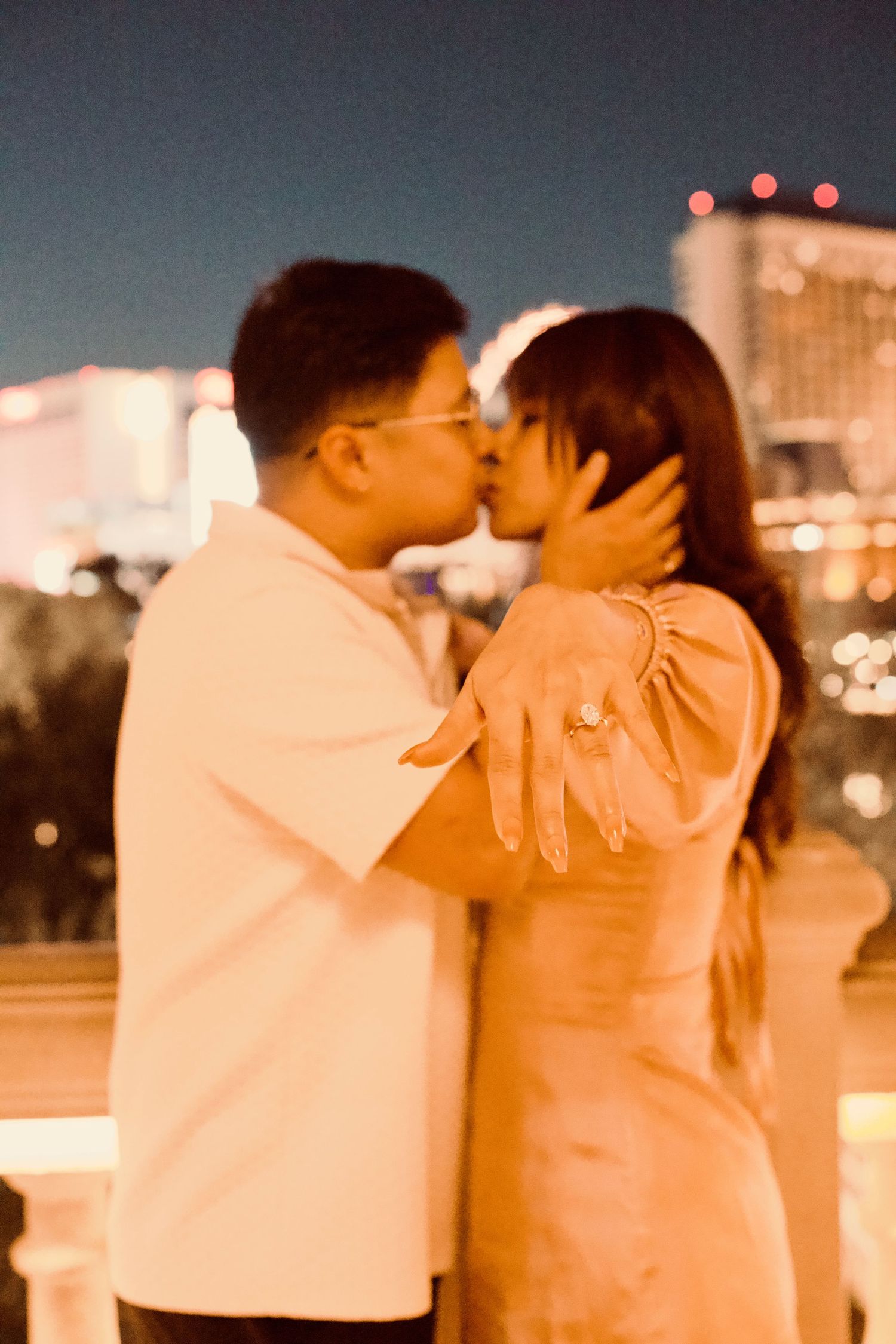 Couple sharing a romantic kiss at night with city lights bokeh background, woman displaying engagement ring.