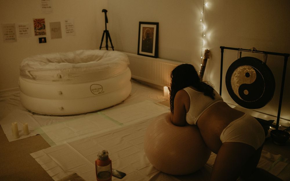 Pregnant woman kneeling on exercise ball near inflatable birth pool and gong in softly lit birth room.