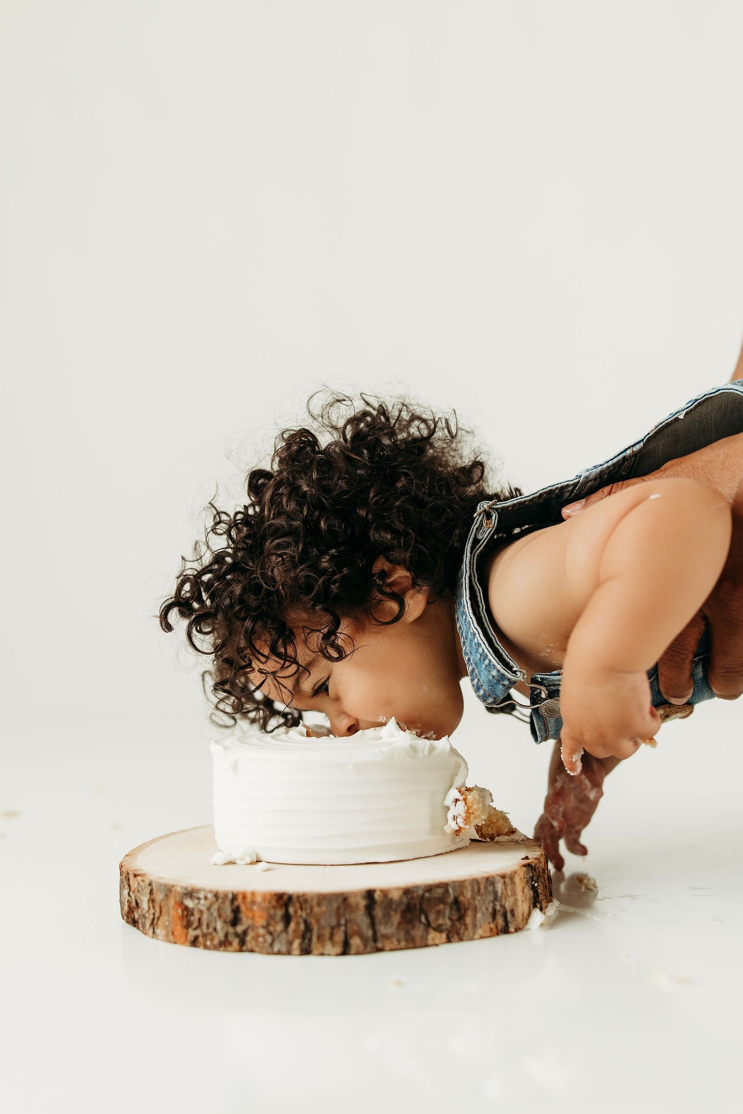 Curly-haired toddler in denim overalls smashing face into white birthday cake on rustic wood slice.