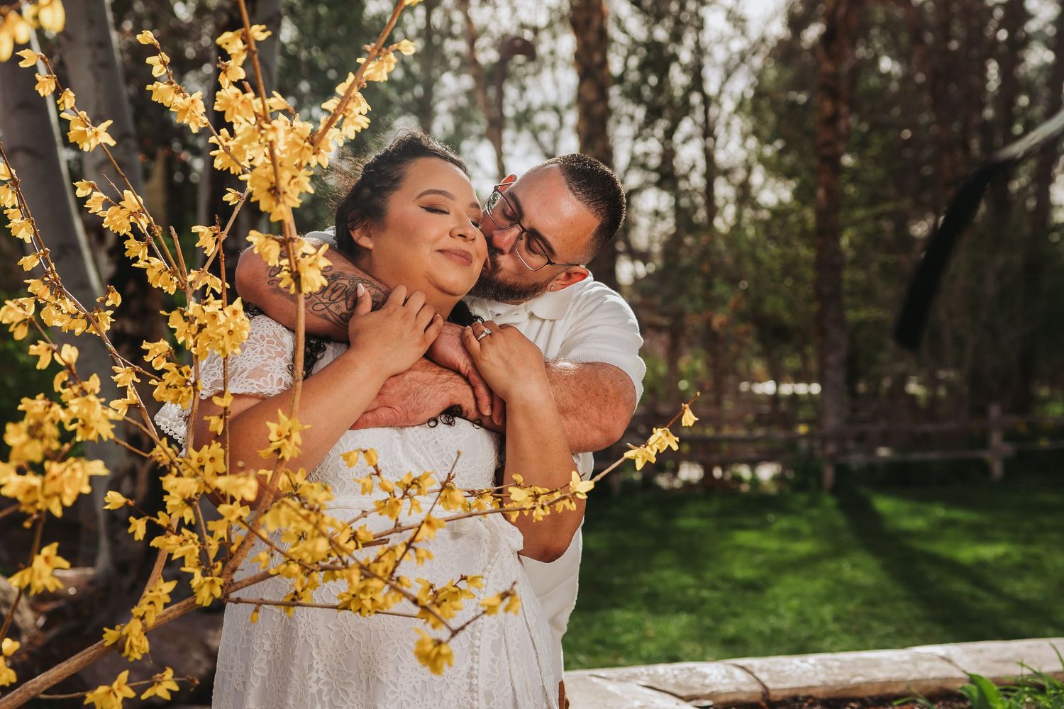 Couple embracing outdoors near yellow blooming forsythia bush in a lush green garden setting.