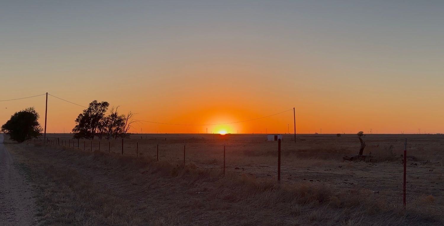A stunning sunset over a flat rural landscape with fence posts and sparse trees silhouetted against a vivid orange sky.