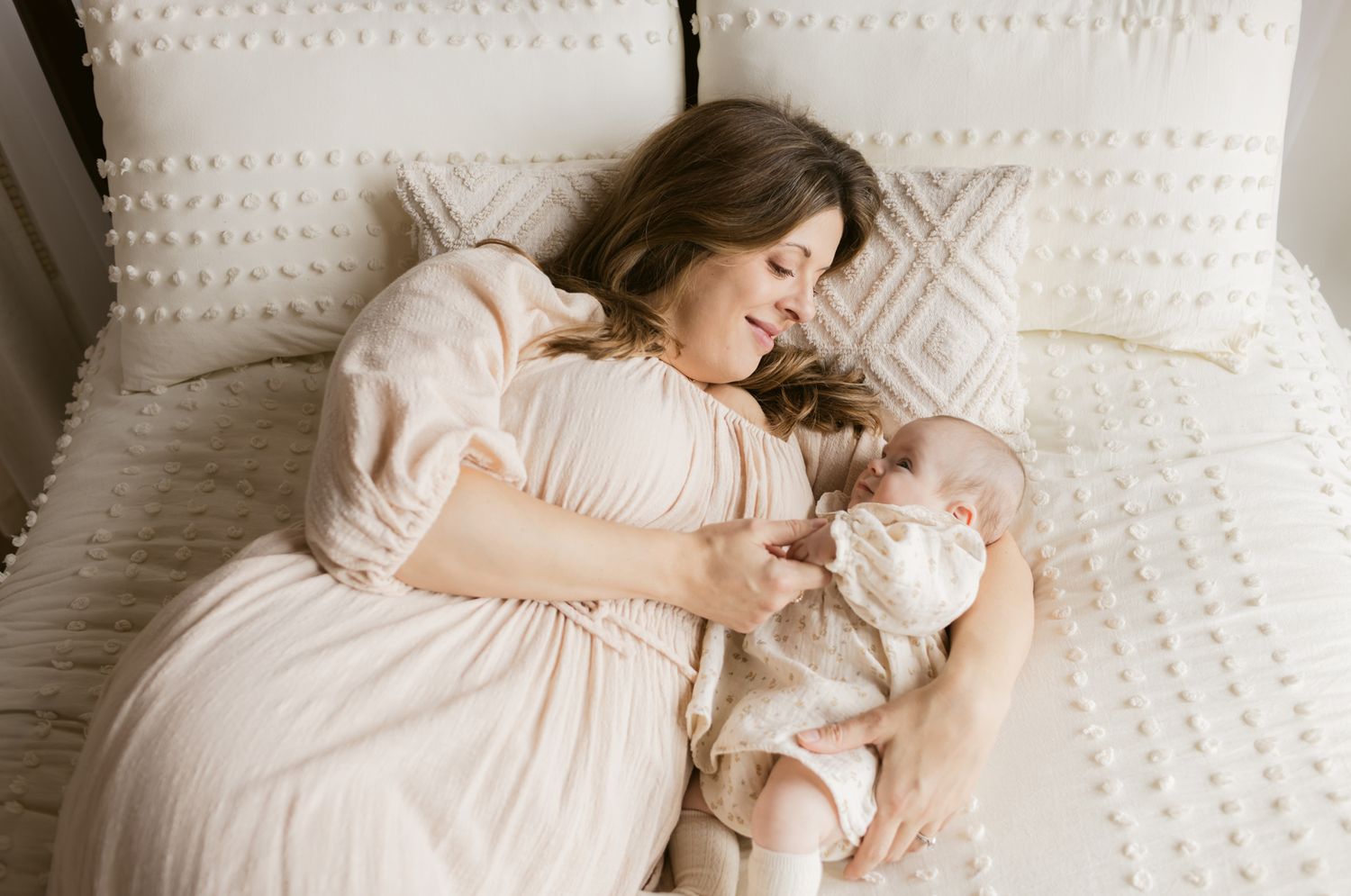 Mother with brown hair wears a cream dress cuddling newborn girl in a floral cream dress on a cream comforter. North Andover newborn photos