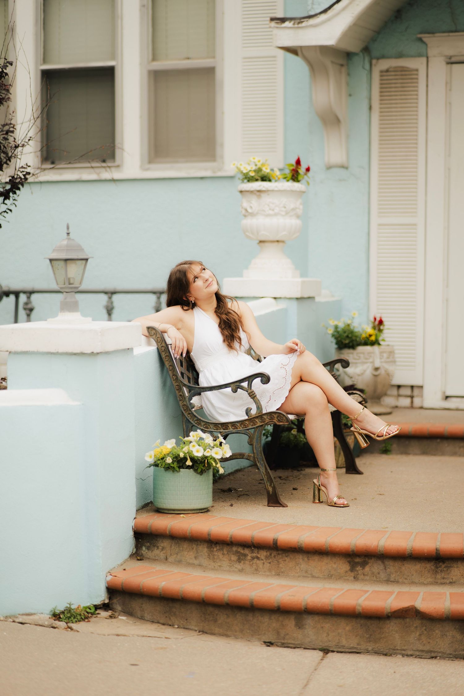 Young woman in white dress relaxing on ornate bench outside a charming light blue Victorian home.
