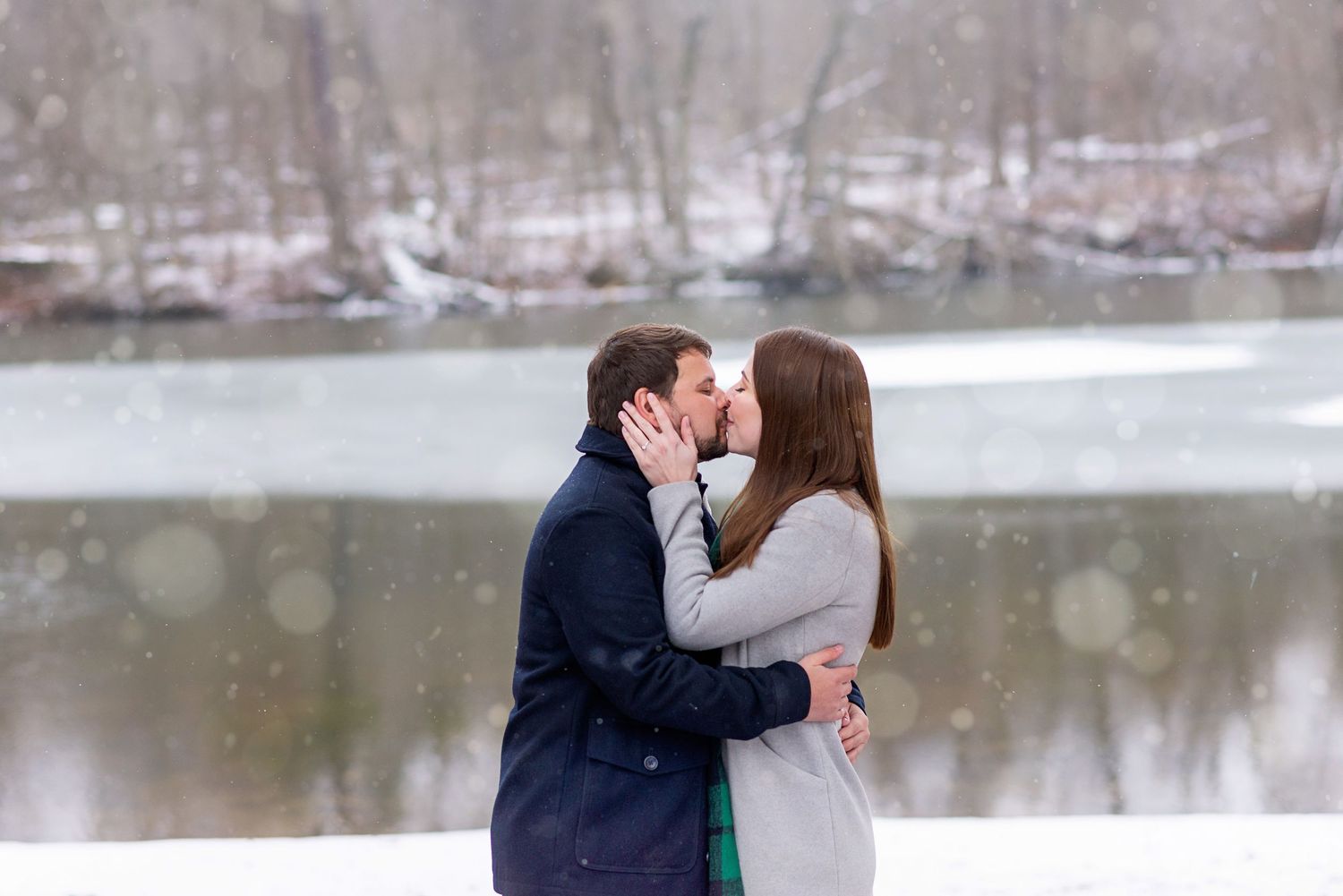 Couple sharing a romantic kiss on a snow-covered dock beside a frozen lake in winter.