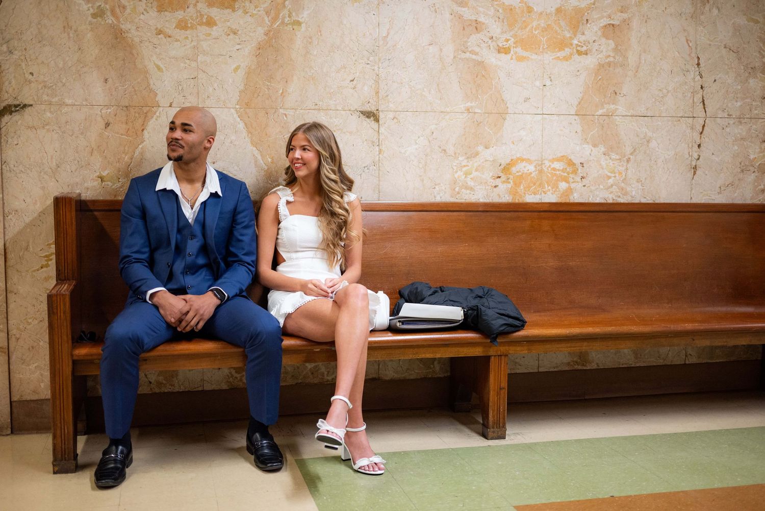Couple in wedding attire sitting together on wooden bench in front of the marble wall outside of a courtroom in Kansas City Missouri