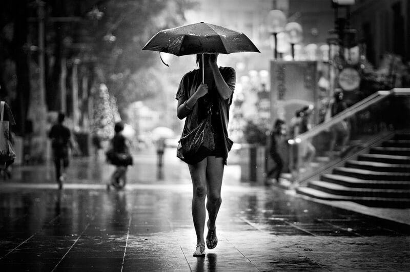 Black and white photo of a woman walking alone in heavy rain holding an umbrella on a busy city street.