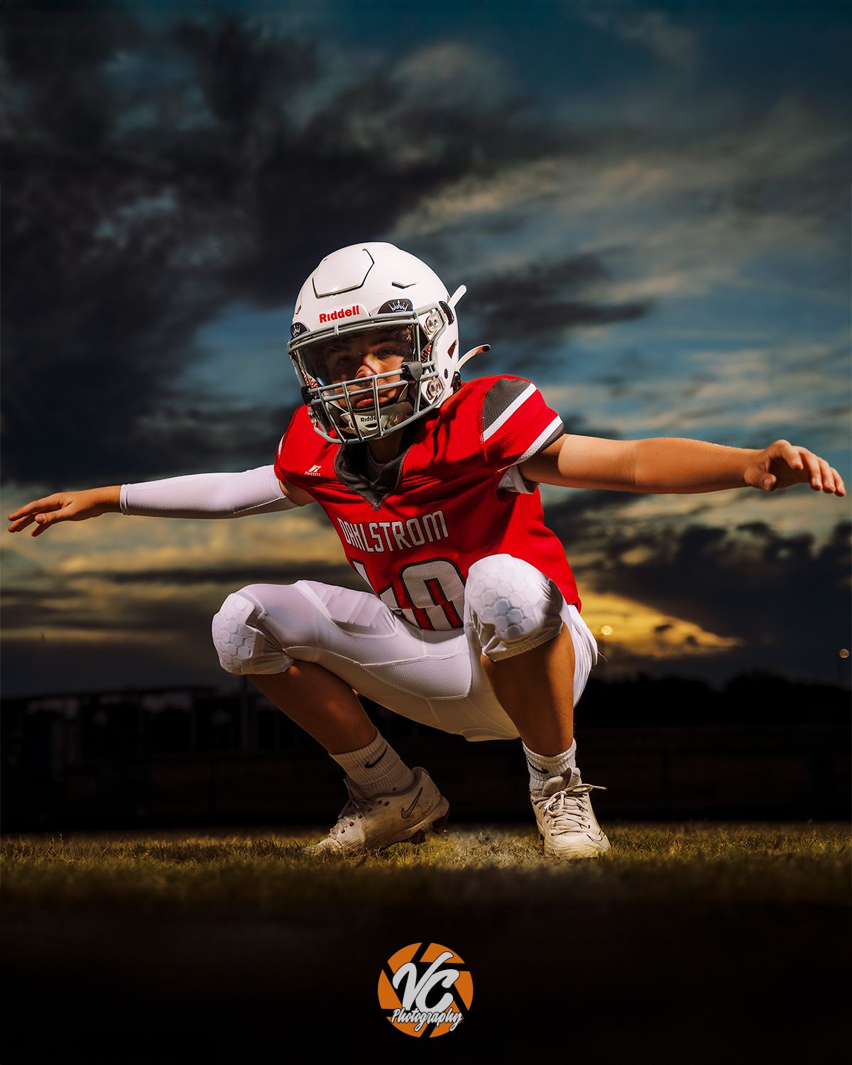 Football player in red jersey and white helmet crouches dynamically on field under dramatic cloudy sky.