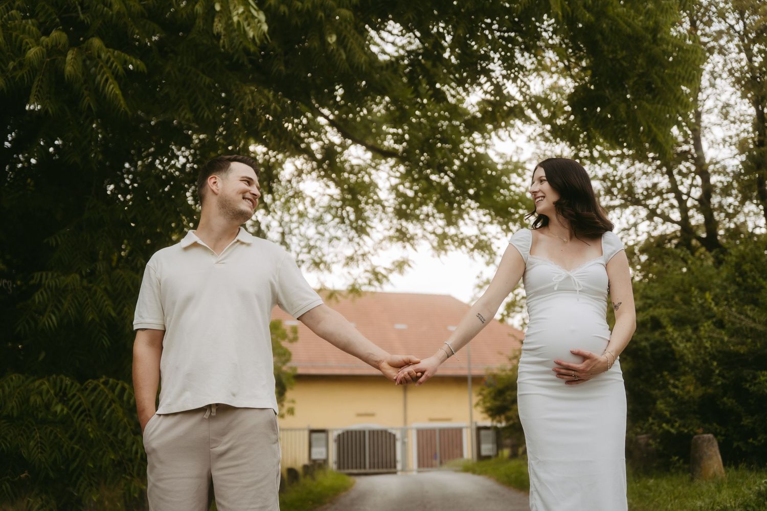 Pregnant couple holding hands outdoors, smiling at each other in front of a yellow house surrounded by lush green trees.