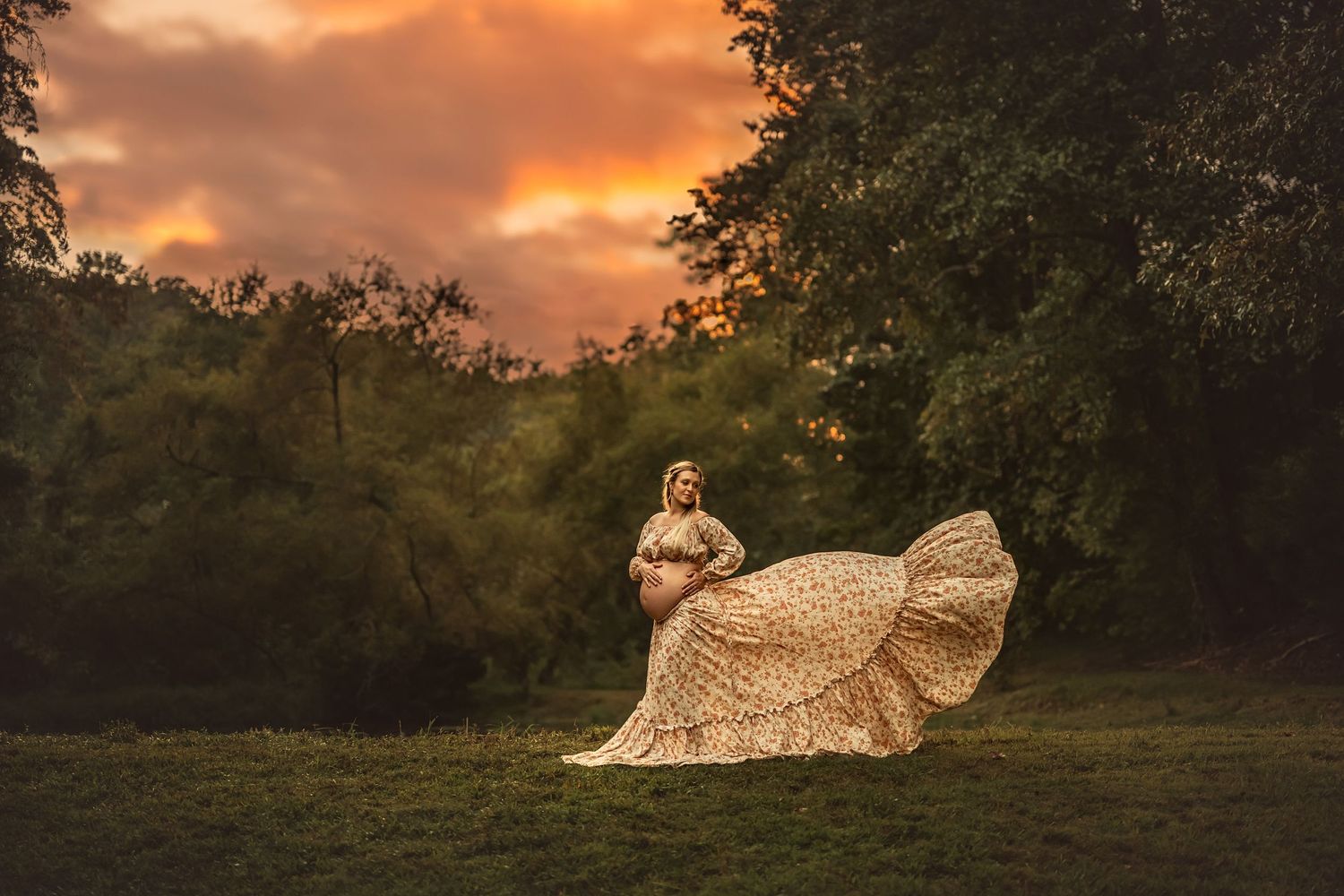 Pregnant woman in flowing floral gown poses outdoors at golden sunset with dramatic orange clouds and lush green trees.