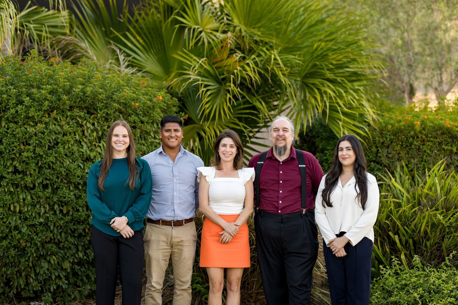 Five professionals smiling outdoors in front of lush green palm trees and tropical foliage on a sunny day.