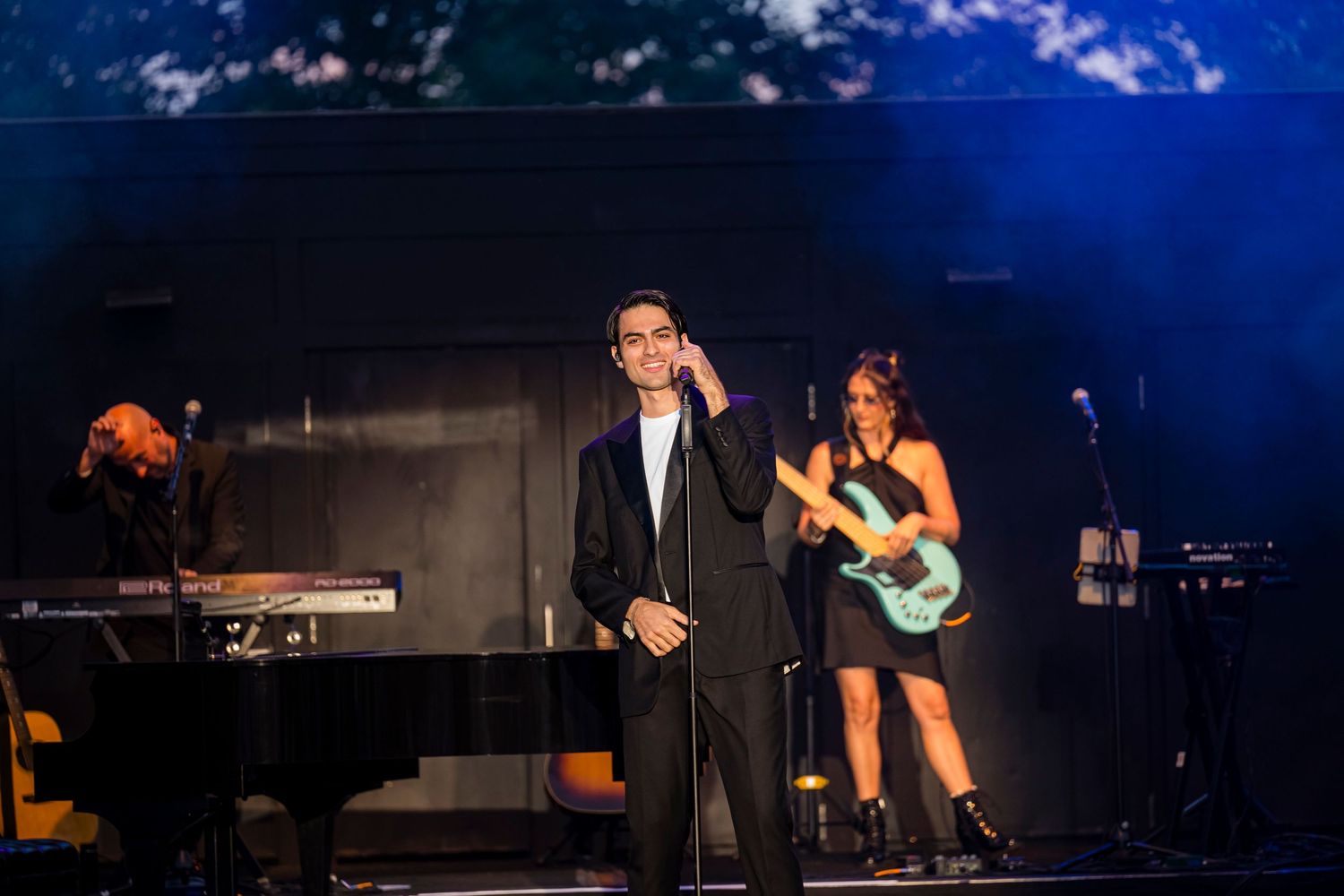 Male performer in black suit on stage with guitarist and keyboardist performing live concert music.