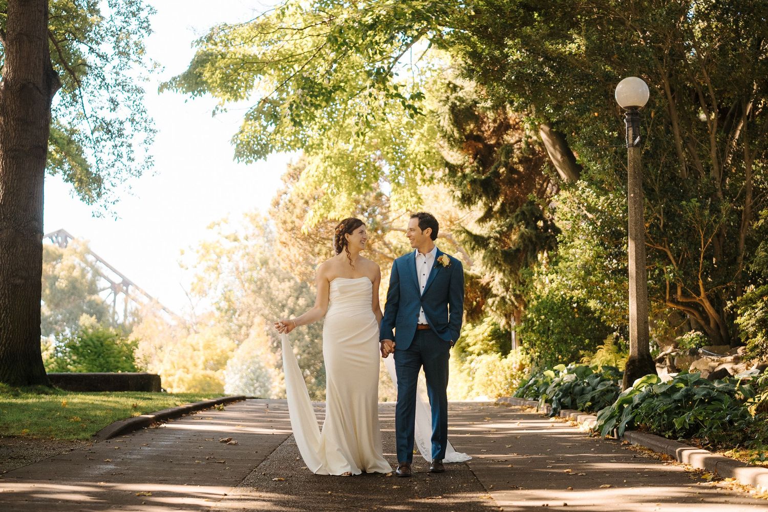 Newlywed couple walks along a tree-lined path with glowing sunlight filtering through the autumn foliage.