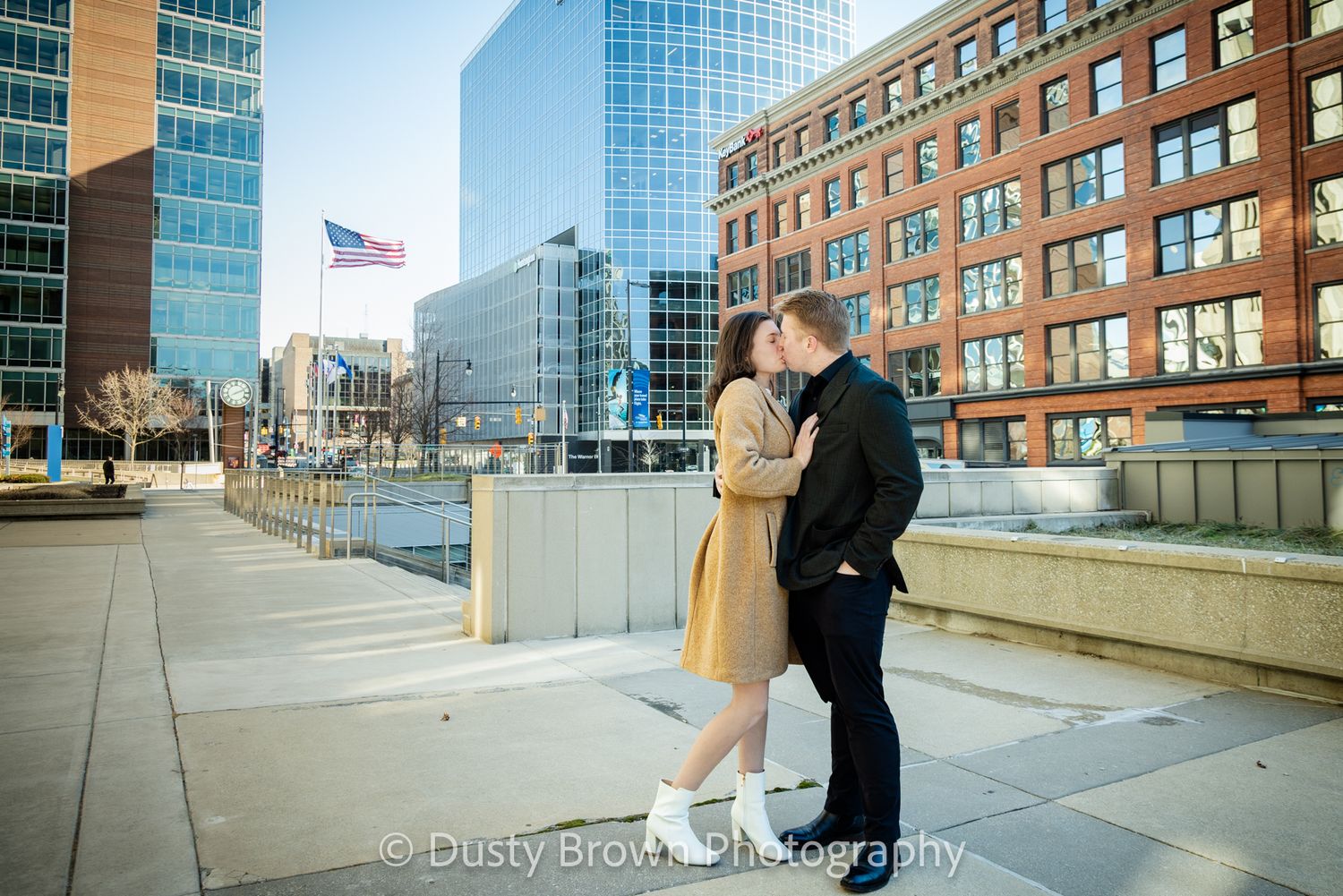 A couple embraces on a rooftop terrace with modern city buildings and glass towers in the background.Grand Rapids Engagement Photographer